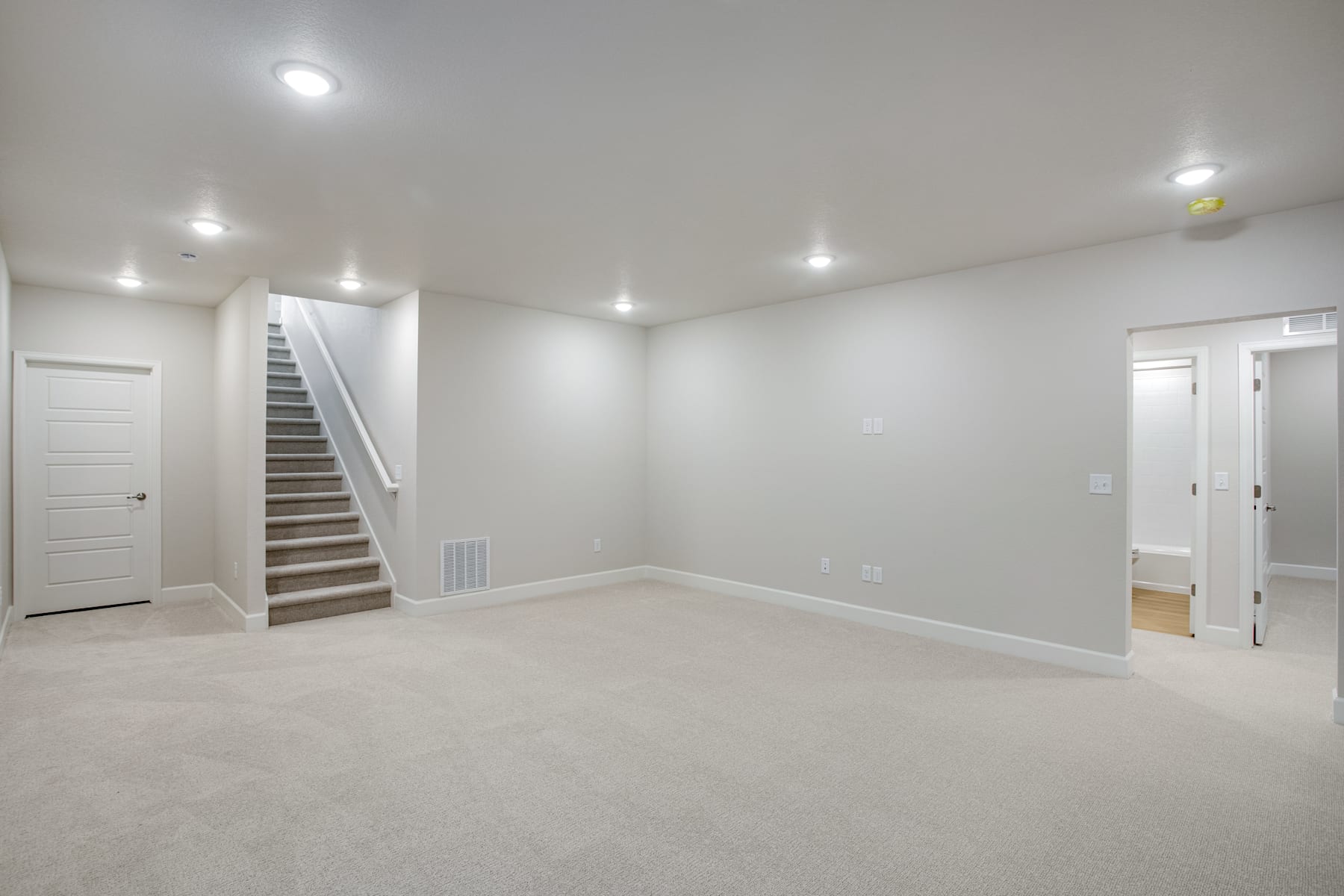 A spacious, well-lit room with a staircase leading to an upper level, and a doorway leading to another room. The room has a clean, minimalist design with white walls and a light-colored carpet.