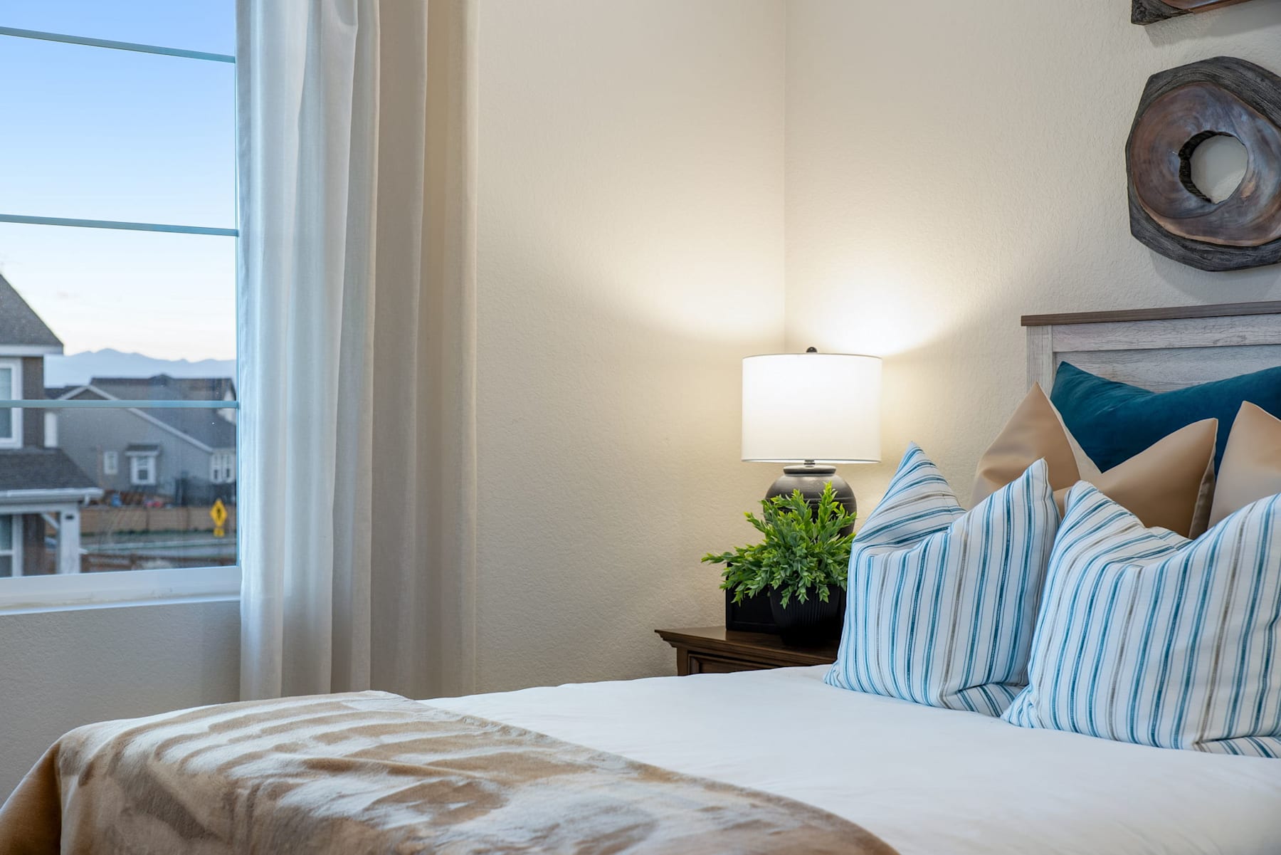 A cozy and inviting bedroom with a large window, a neatly made bed with striped and teal pillows, and a potted plant adding a touch of greenery to the space.