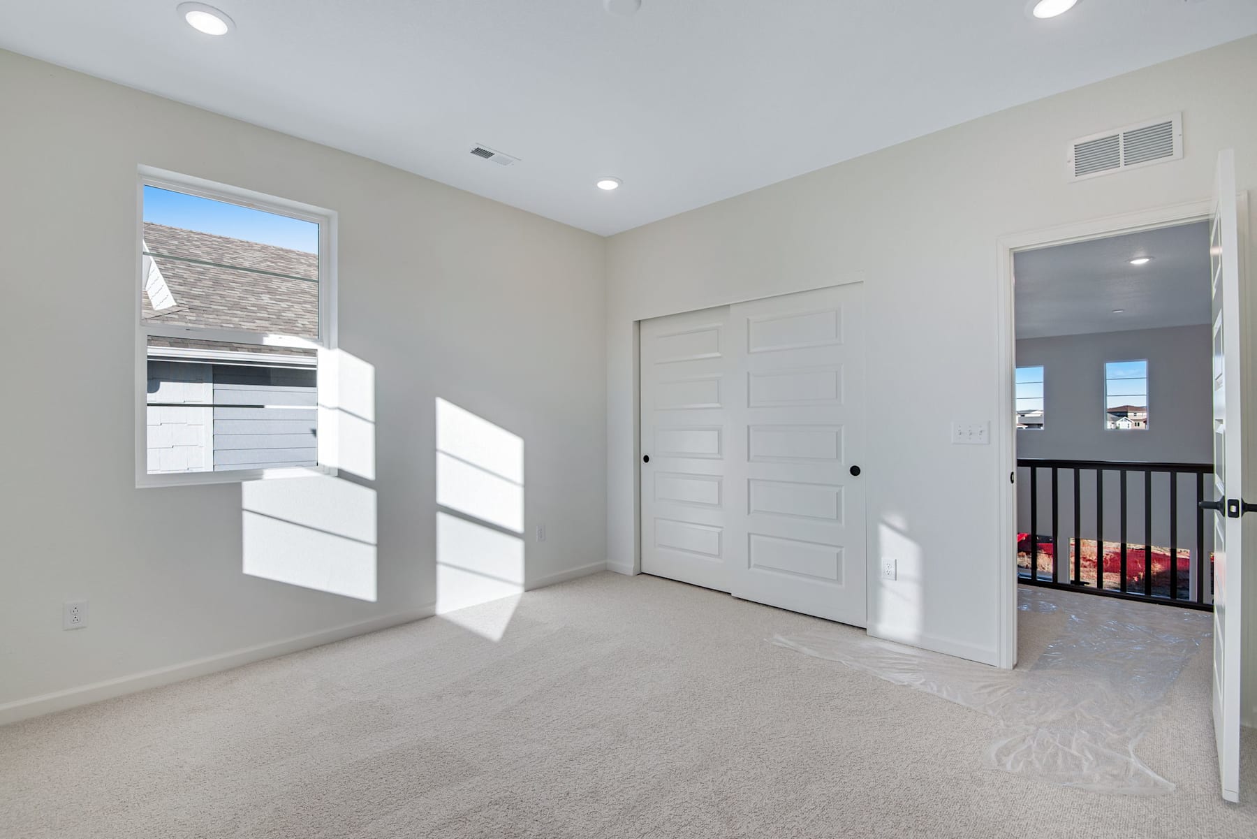 A spacious, well-lit room with white walls, a carpeted floor, and a closed door leading to another area. The room appears to be part of a larger residential space, with natural light streaming in through the windows.