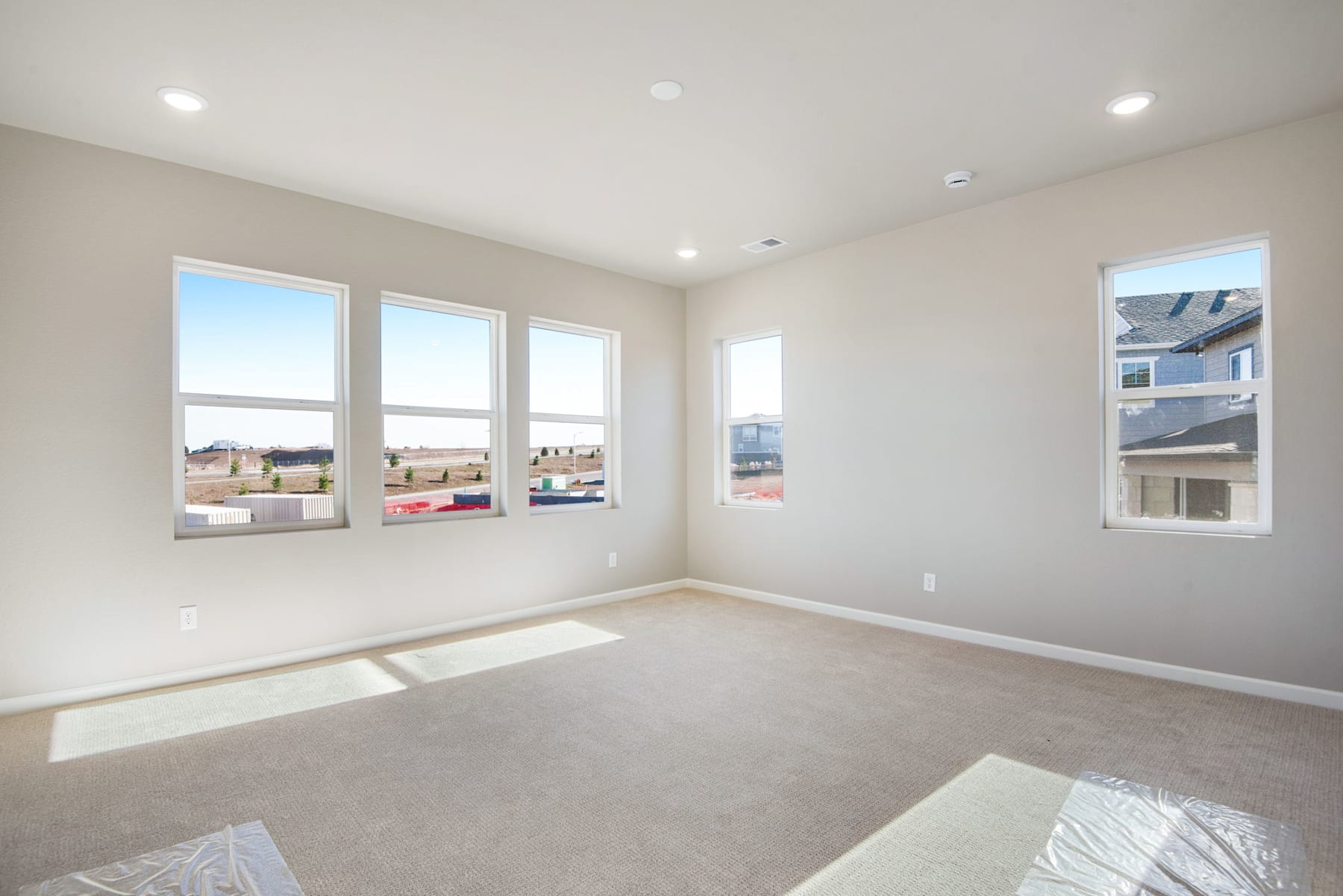 A spacious, well-lit room with large windows overlooking a residential neighborhood, featuring neutral-toned walls, plush carpeting, and recessed lighting.