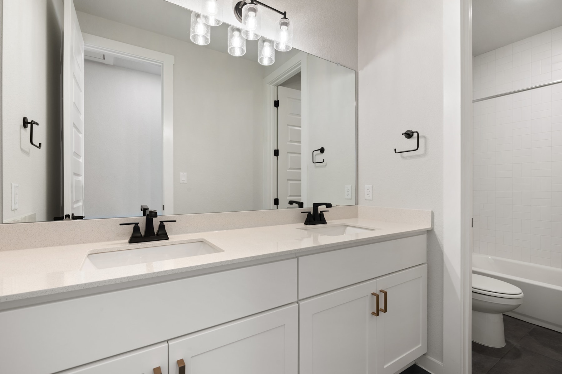A modern, minimalist bathroom with a white vanity, dual sinks, and a large mirror above, illuminated by a sleek light fixture.