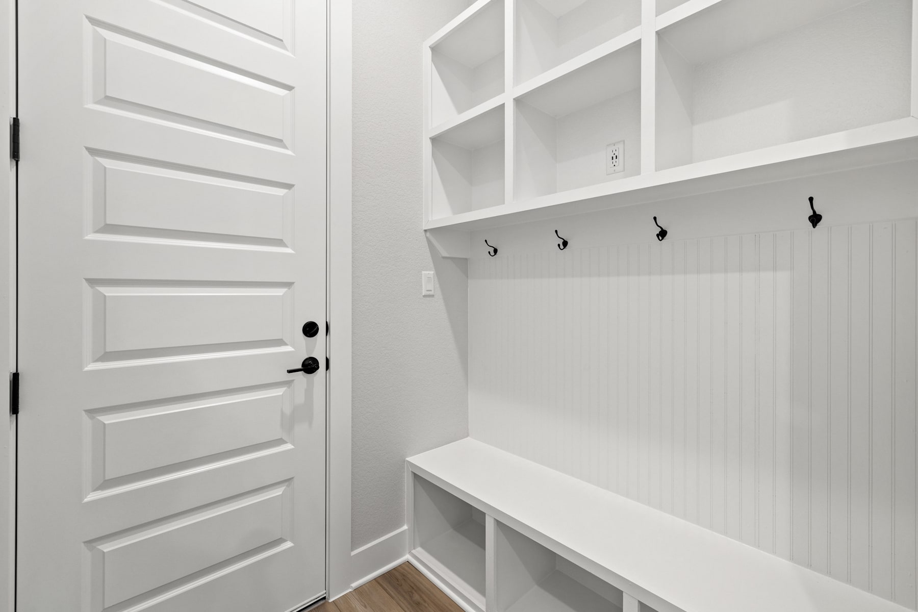 A bright, white mudroom with a wooden bench, hooks, and shelves, leading to a closed door.