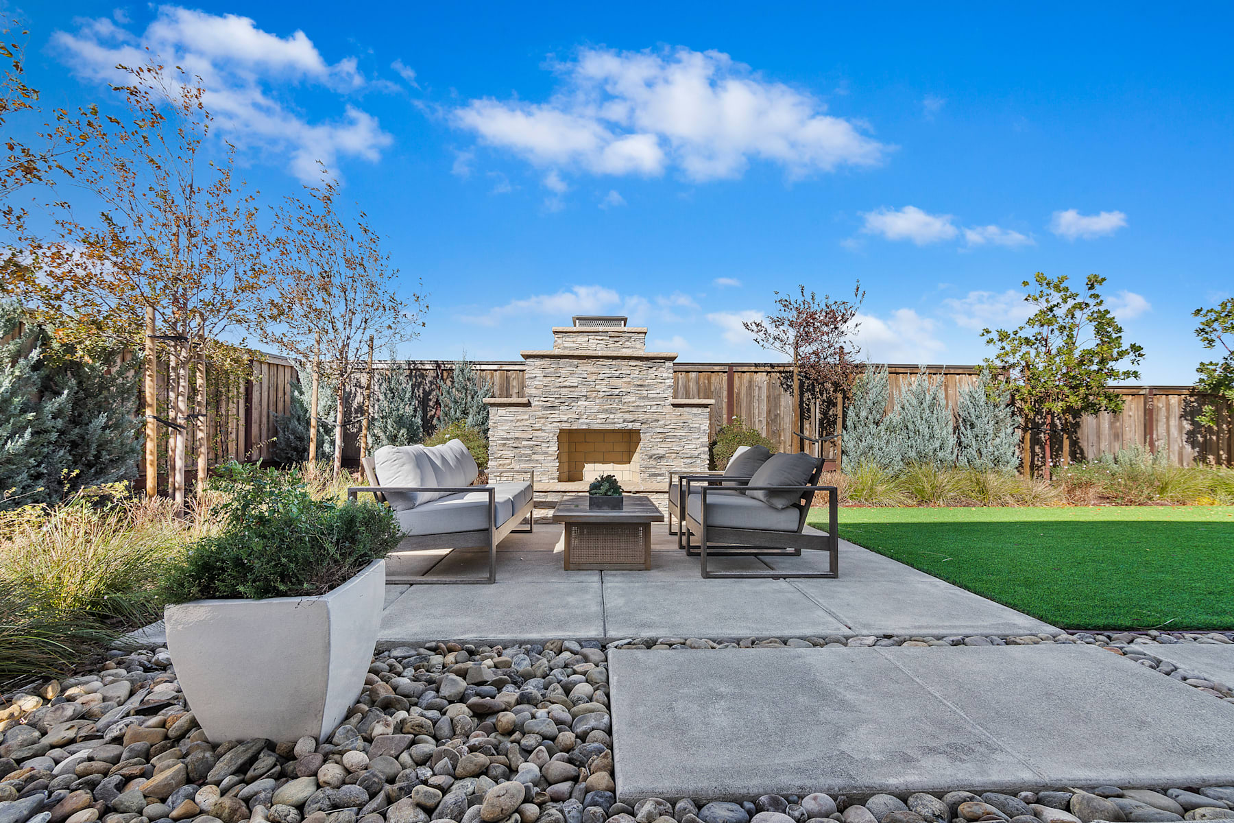 A modern outdoor living space with a stone fireplace, comfortable seating, and a lush, landscaped backyard under a bright, blue sky with fluffy clouds.