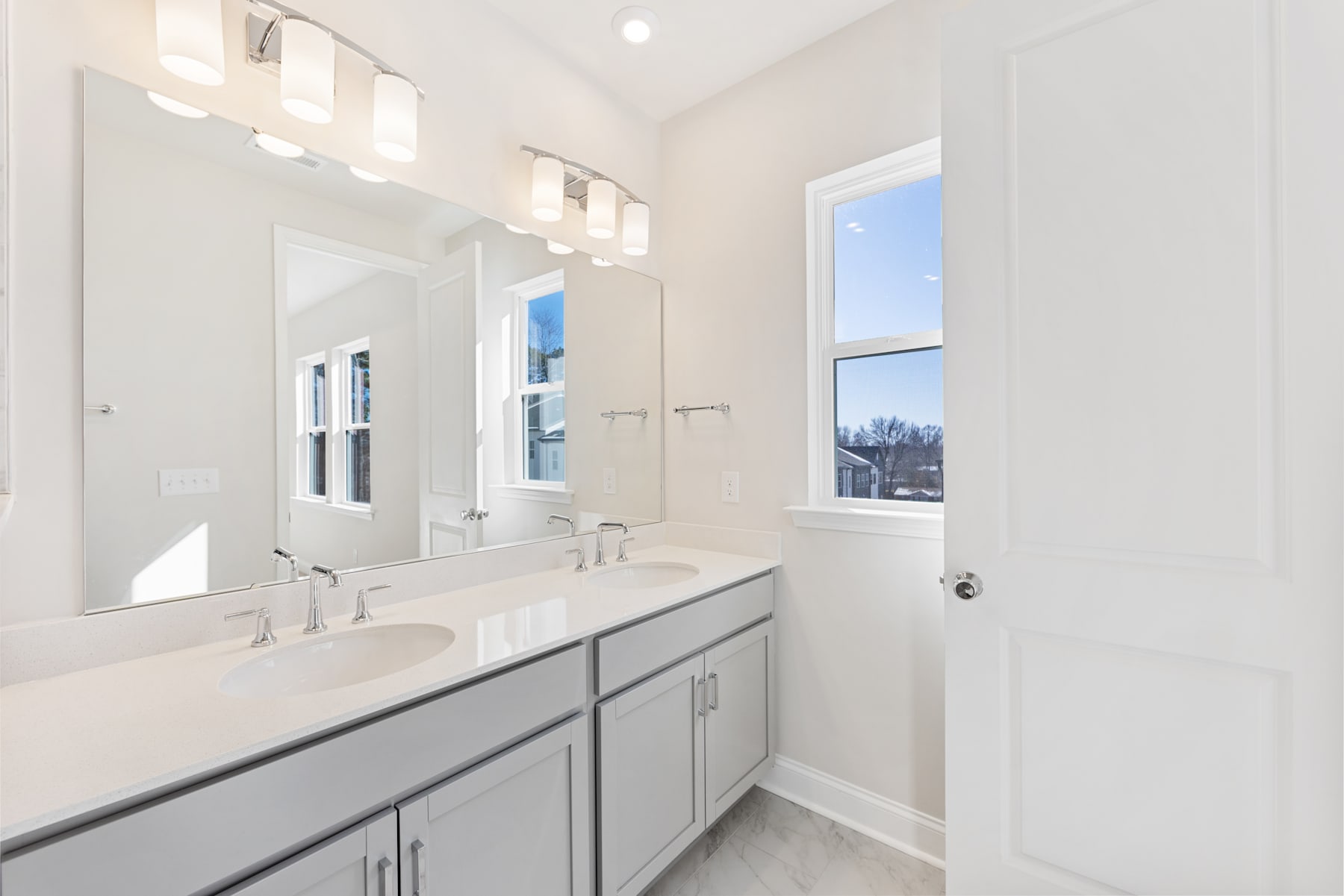 A modern, well-lit bathroom with a double vanity, white walls, and a large window overlooking the outdoors.