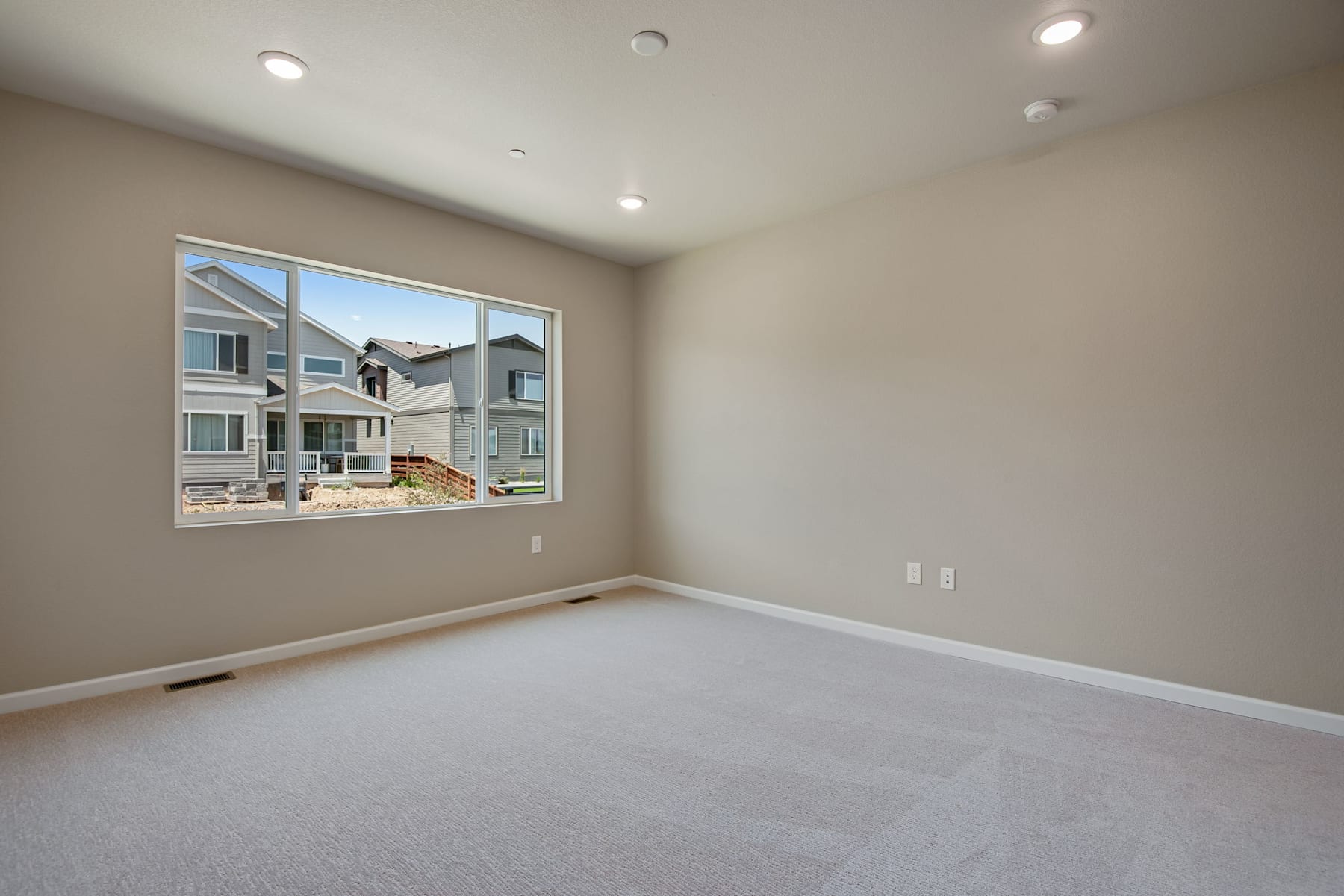 An empty room with a large window overlooking a residential neighborhood, featuring a neutral-colored wall and a carpeted floor.
