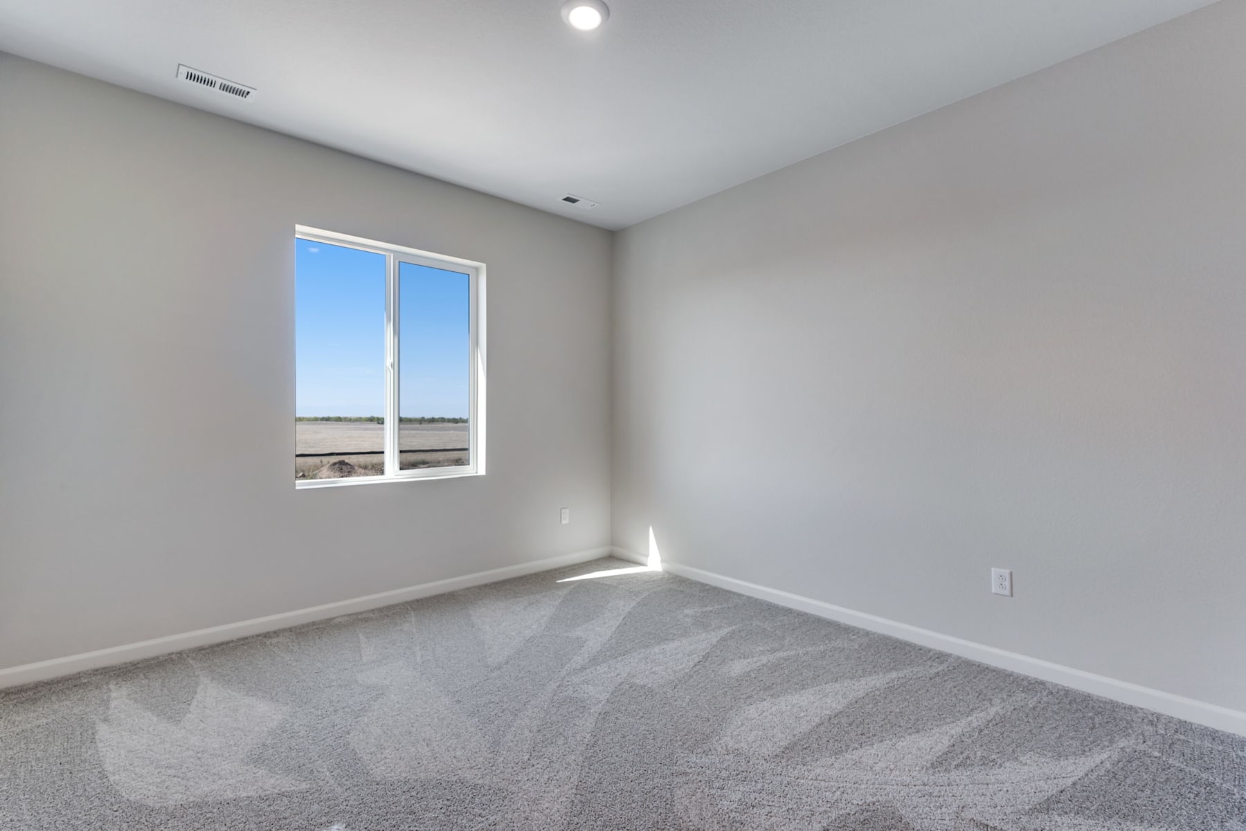 A spacious, well-lit room with a large window overlooking the outdoors, featuring a gray carpeted floor and plain white walls.