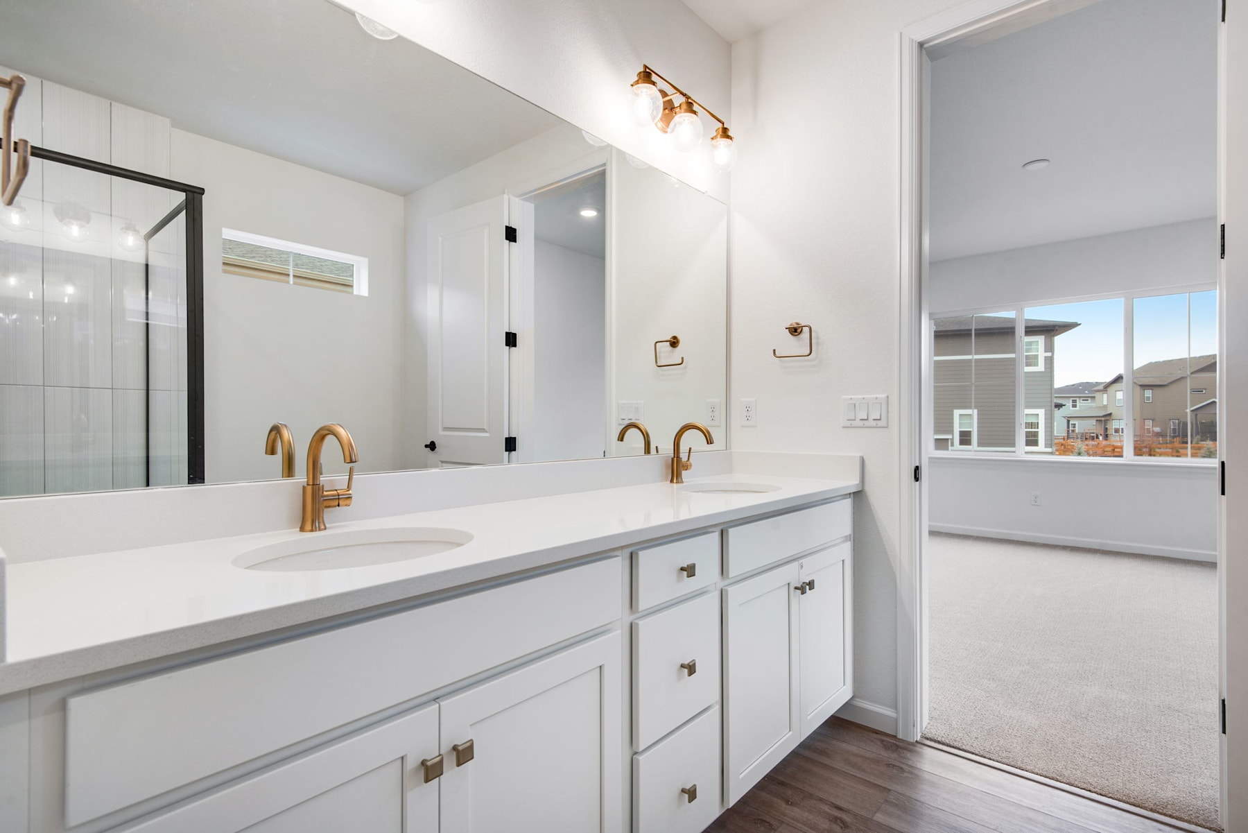A modern, well-lit bathroom with a double vanity, white cabinets, and gold-toned fixtures, set against a backdrop of a large window overlooking an outdoor scene.