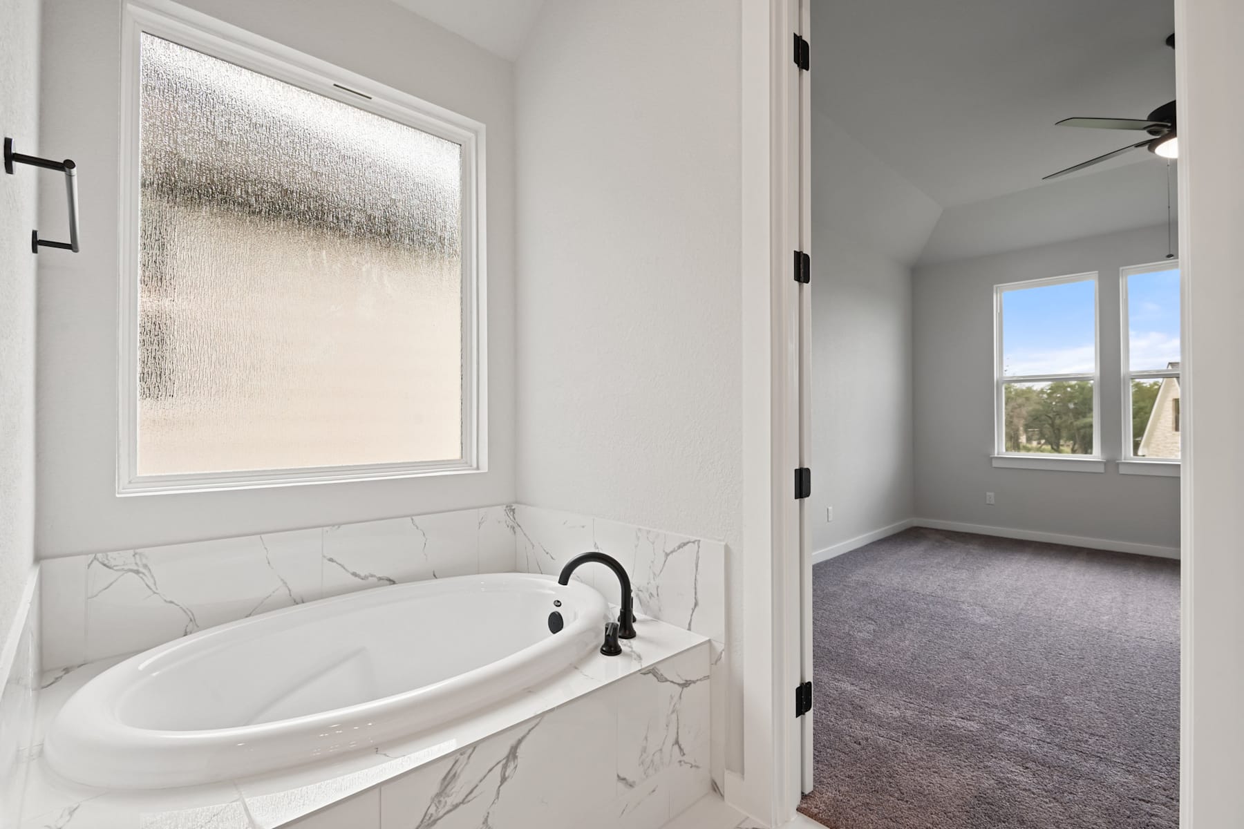 A modern bathroom with a white bathtub and a window with a frosted glass panel, leading to an adjacent room with a carpeted floor and a ceiling fan.