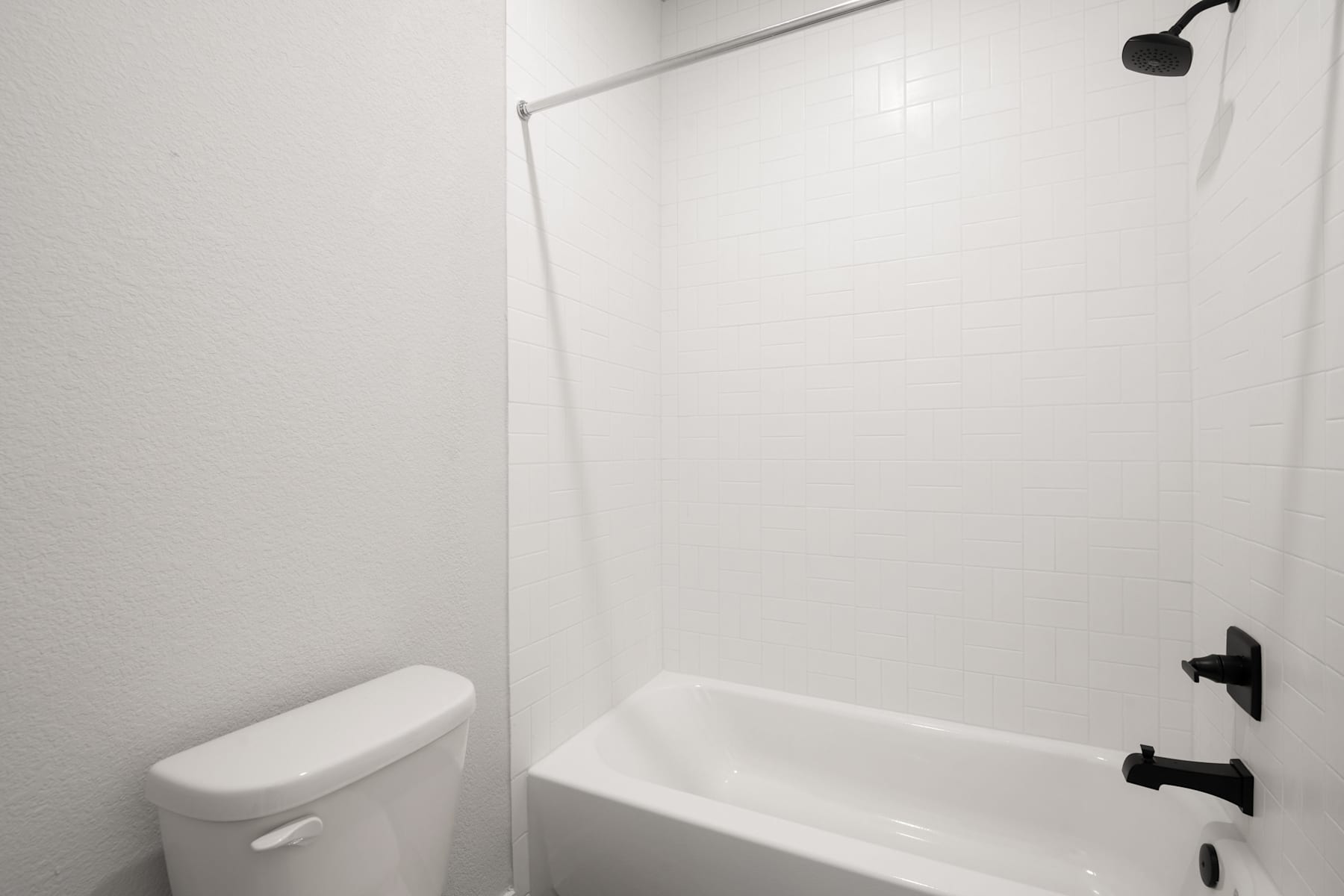 A simple, minimalist bathroom with a white toilet, bathtub, and shower fixtures against a plain white wall.