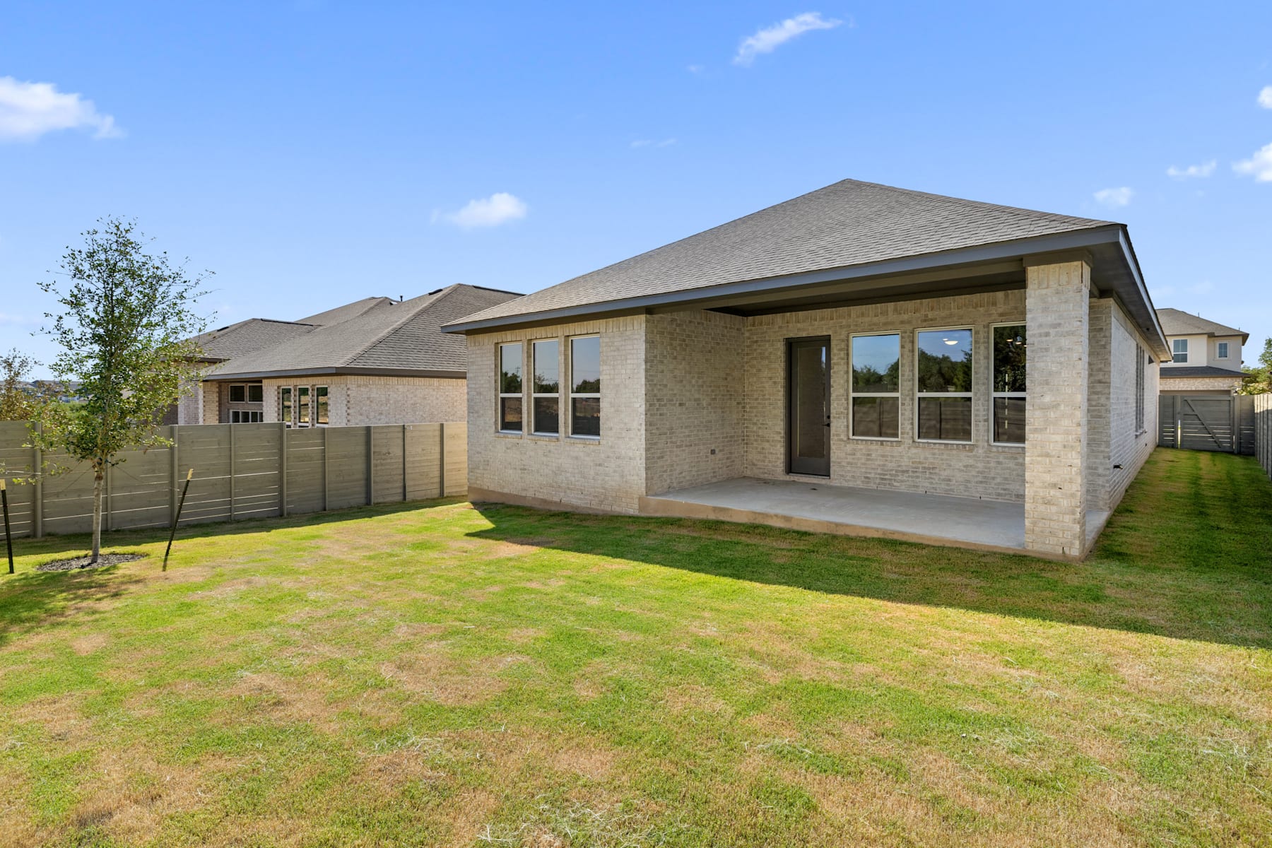 A single-story house with a gray exterior and a slanted roof stands in a grassy yard, surrounded by trees and a clear blue sky.