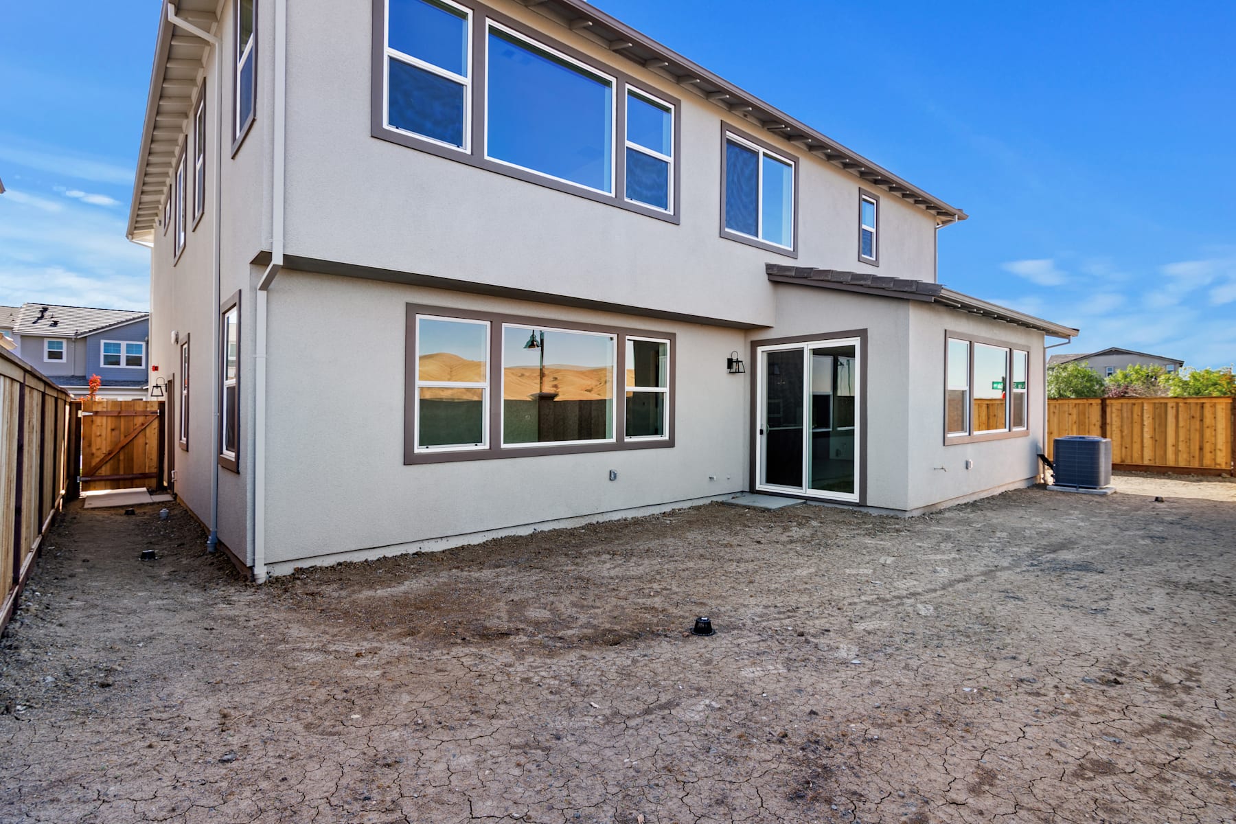 A two-story residential building with a gray exterior, surrounded by a dirt yard and a wooden fence in the foreground, set against a clear blue sky in the background.