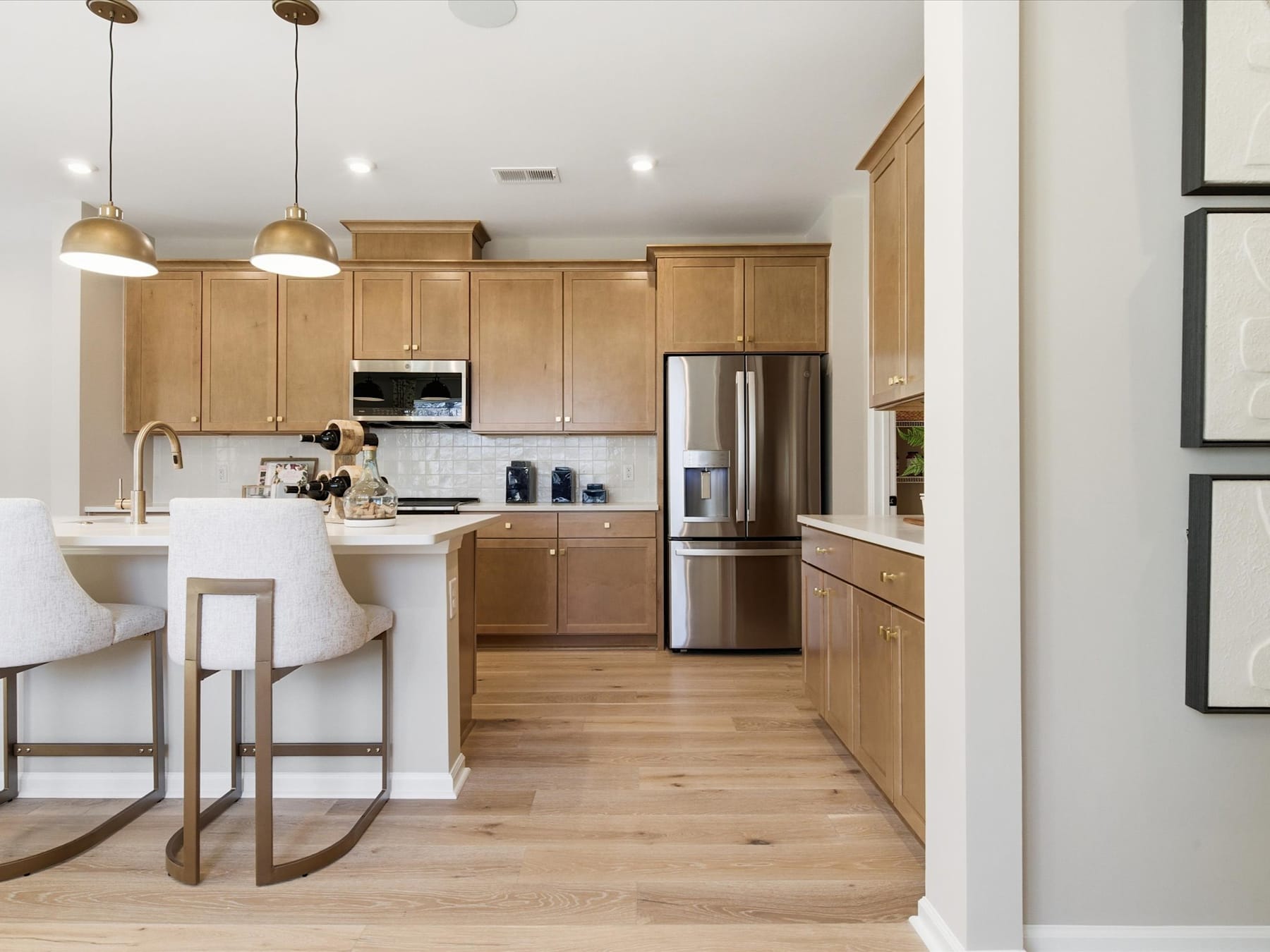 A modern, open-concept kitchen with light wood cabinetry, stainless steel appliances, and a central island with bar stools, set against a backdrop of white walls and hardwood floors.