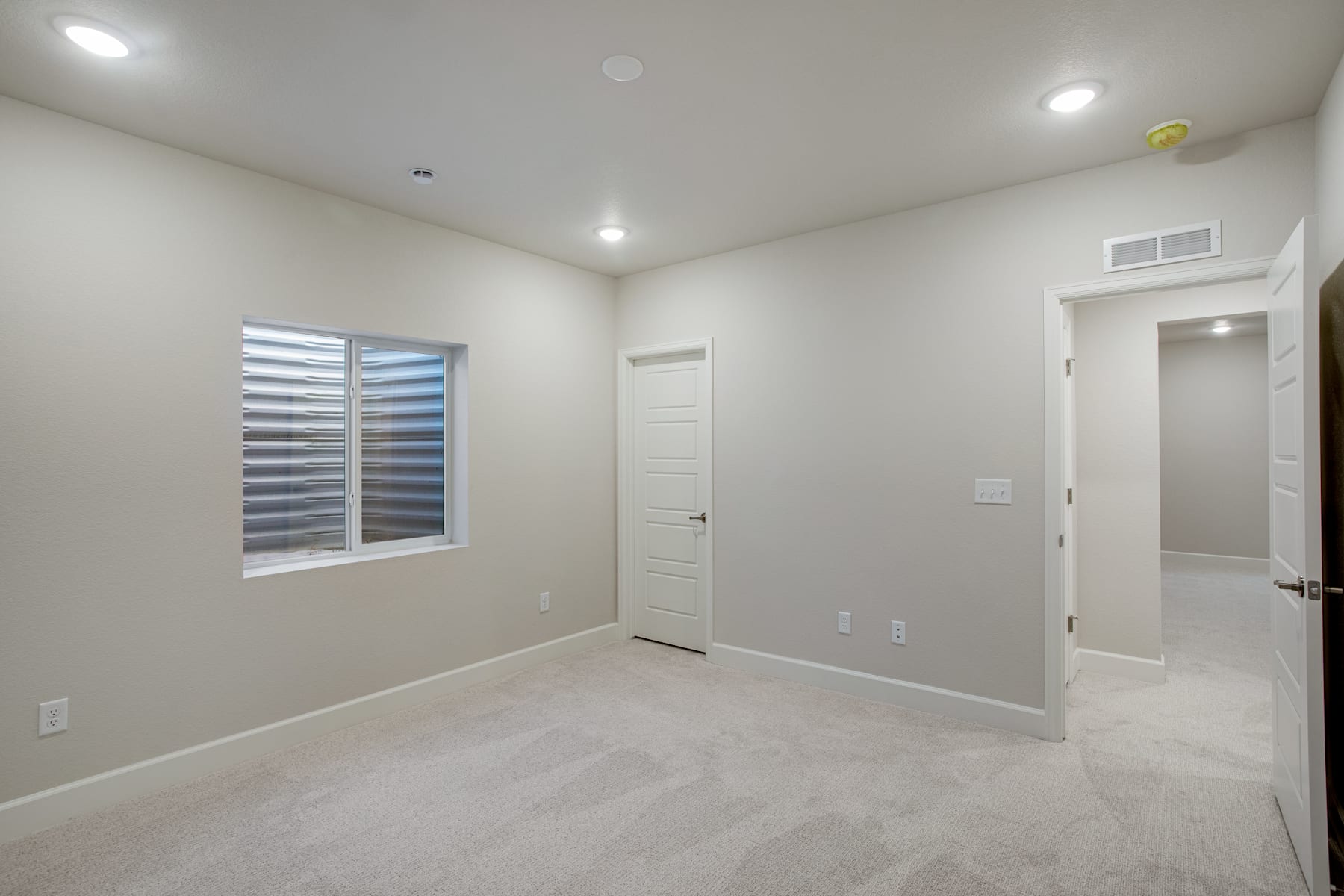 A spacious, well-lit room with a window, a door, and a neutral-colored carpet covering the floor.