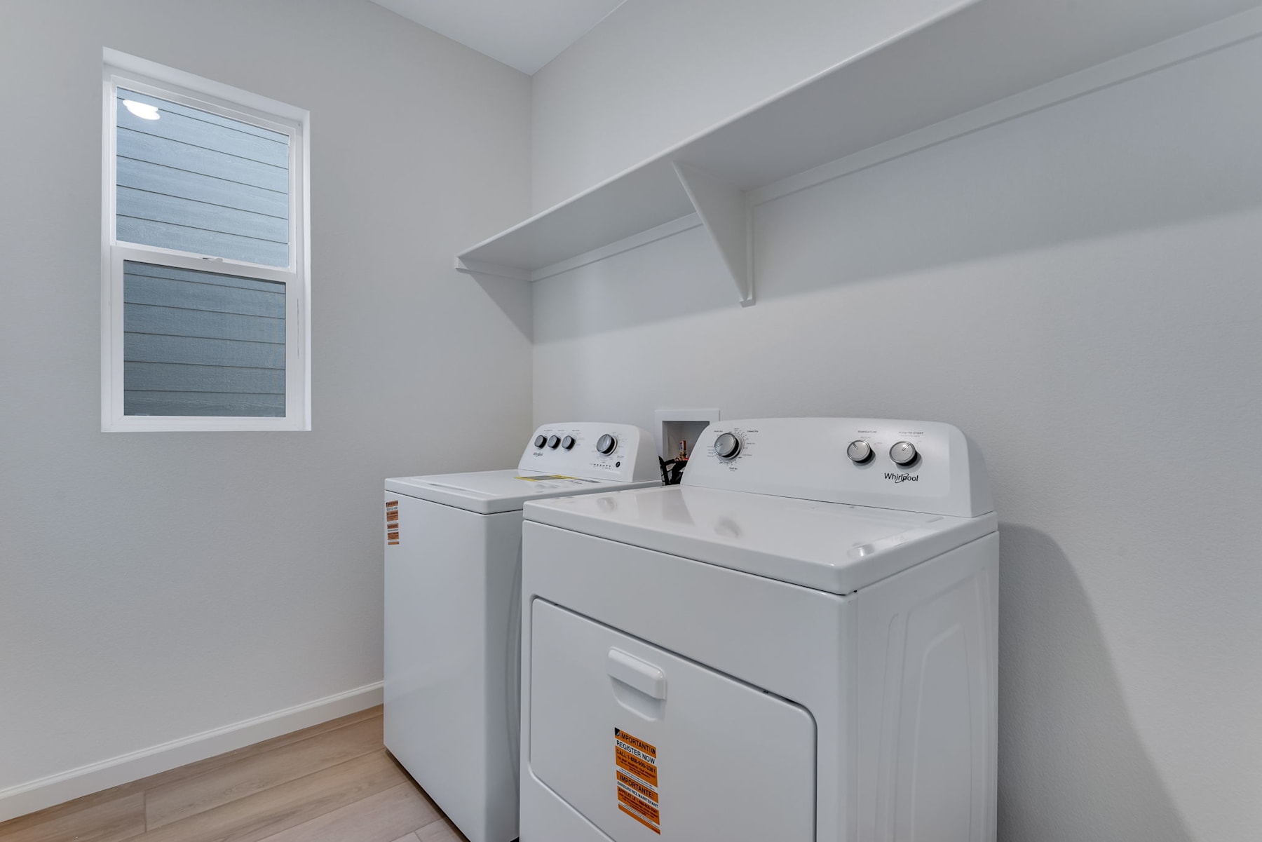 A modern, well-lit laundry room with a washing machine and dryer set against a plain white wall, with a small window providing natural light.