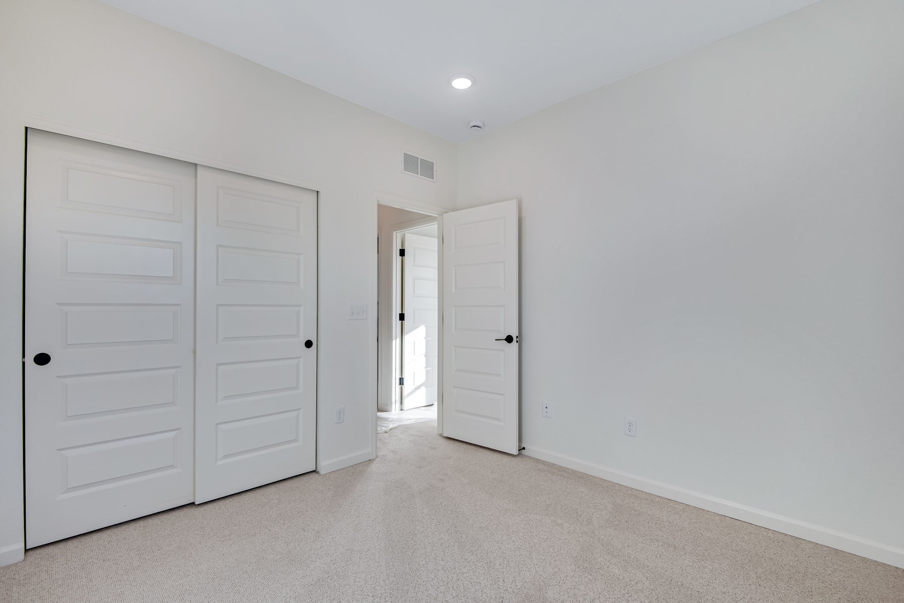 A bright, minimalist bedroom with white walls, light-colored hardwood floors, and two white closet doors in the foreground.