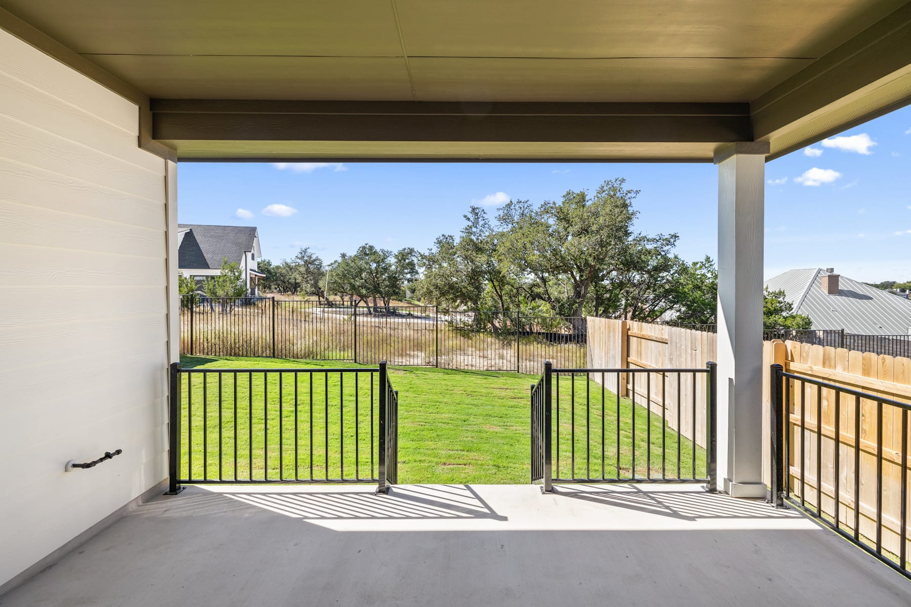 A covered patio with a green lawn and fenced yard in the background, surrounded by trees and a clear blue sky.