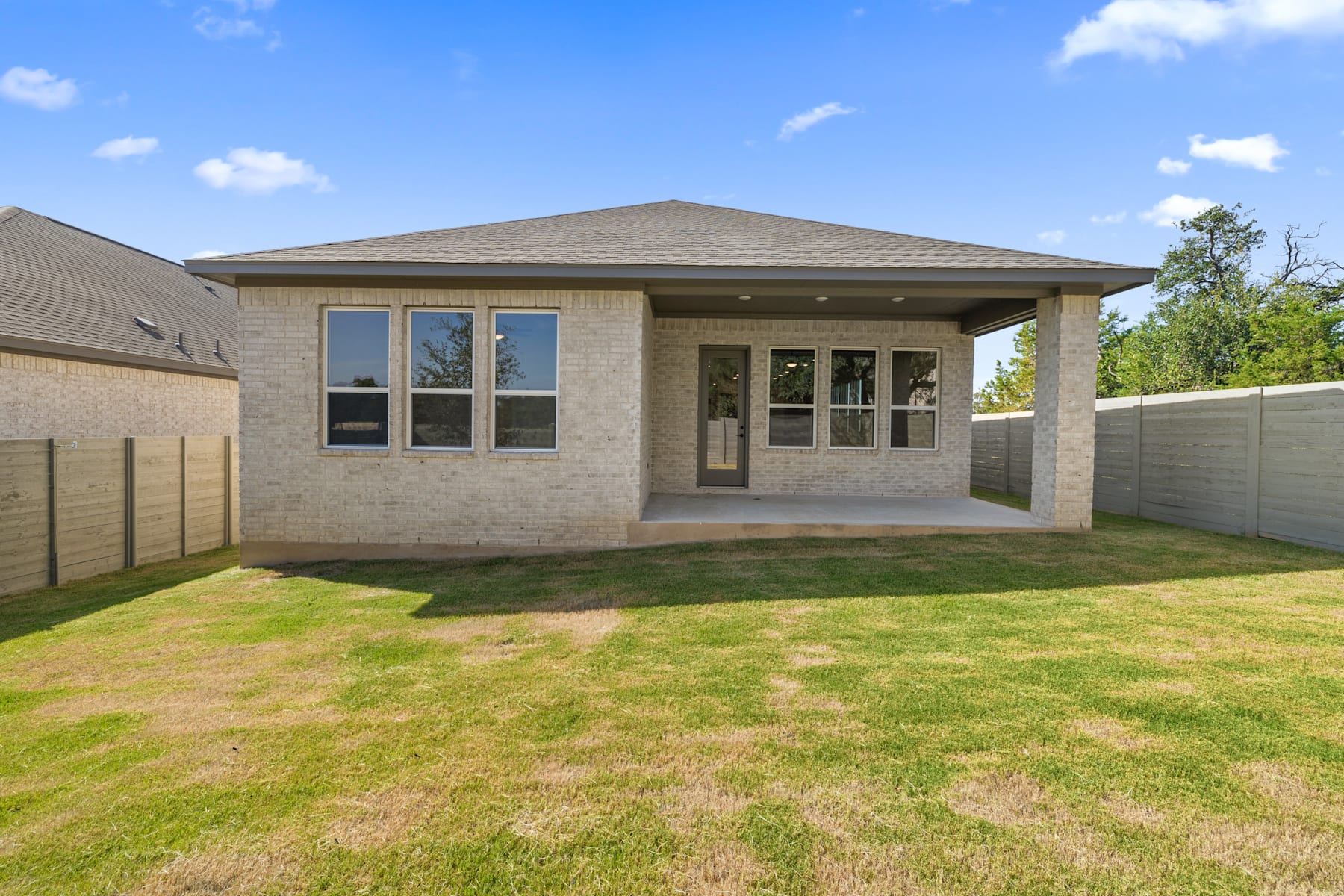 A single-story brick house with a covered porch stands in a grassy yard, surrounded by trees and a clear blue sky with scattered clouds.