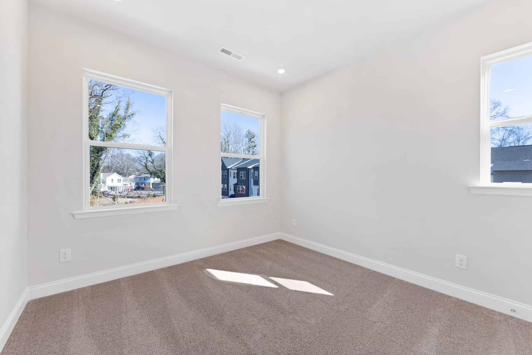 A bright, empty room with large windows overlooking a residential neighborhood, featuring hardwood floors and neutral-colored walls.