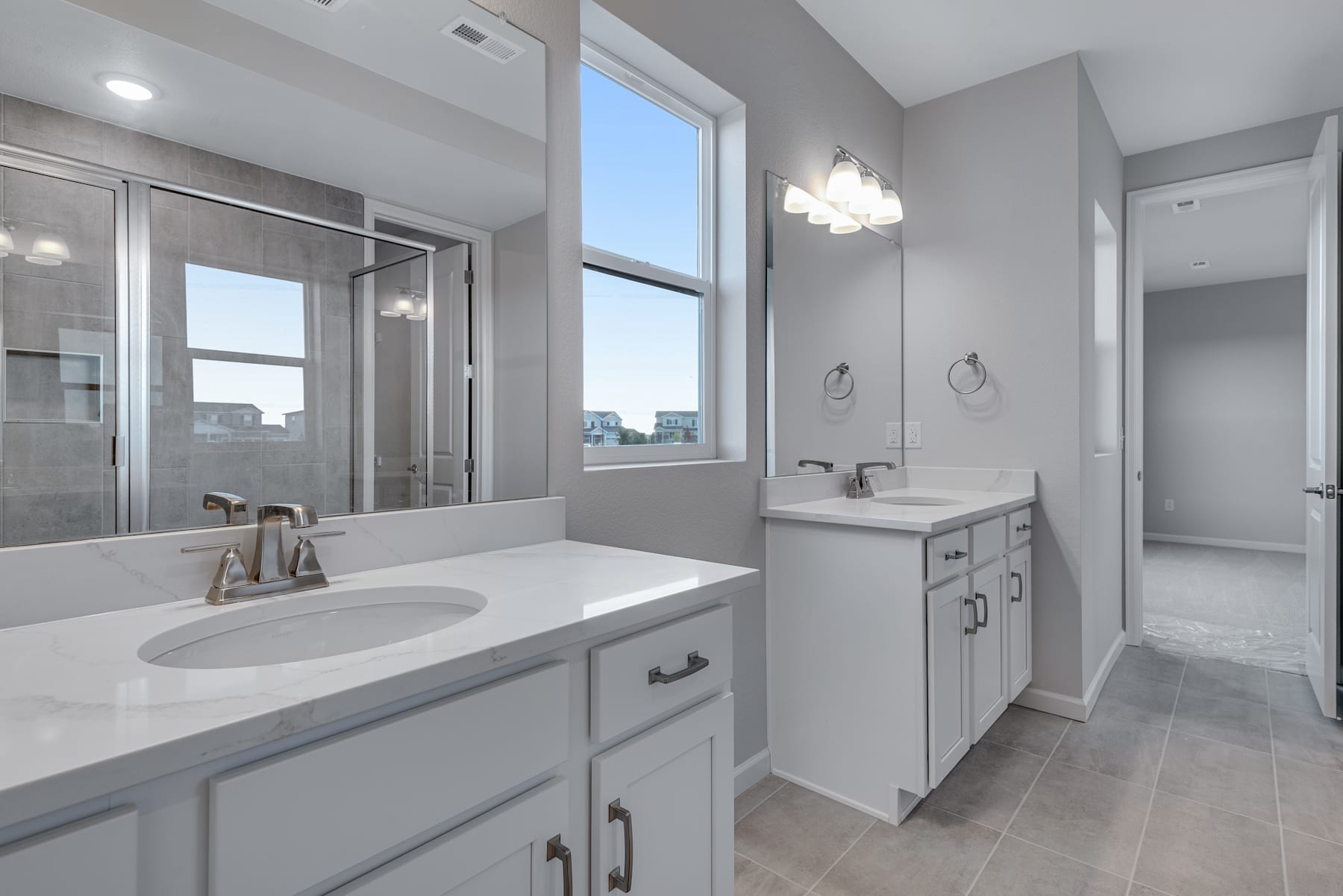 A modern, well-lit bathroom with white vanity, countertop, and cabinets, along with a large window providing natural light and a view of the cityscape in the background.