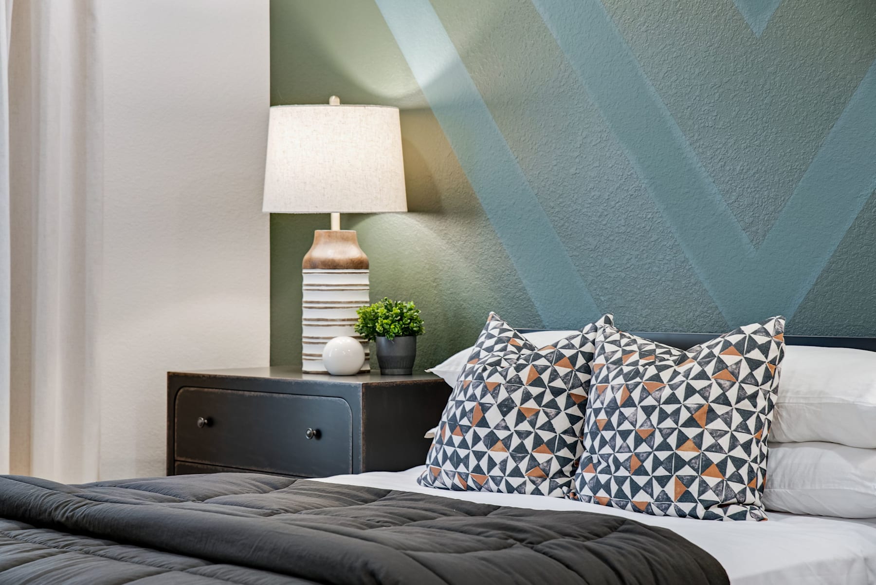 A cozy bedroom with a patterned throw pillow on a gray bedspread, a wooden nightstand with a lamp, and a potted plant, set against a blue and white geometric-patterned wall.