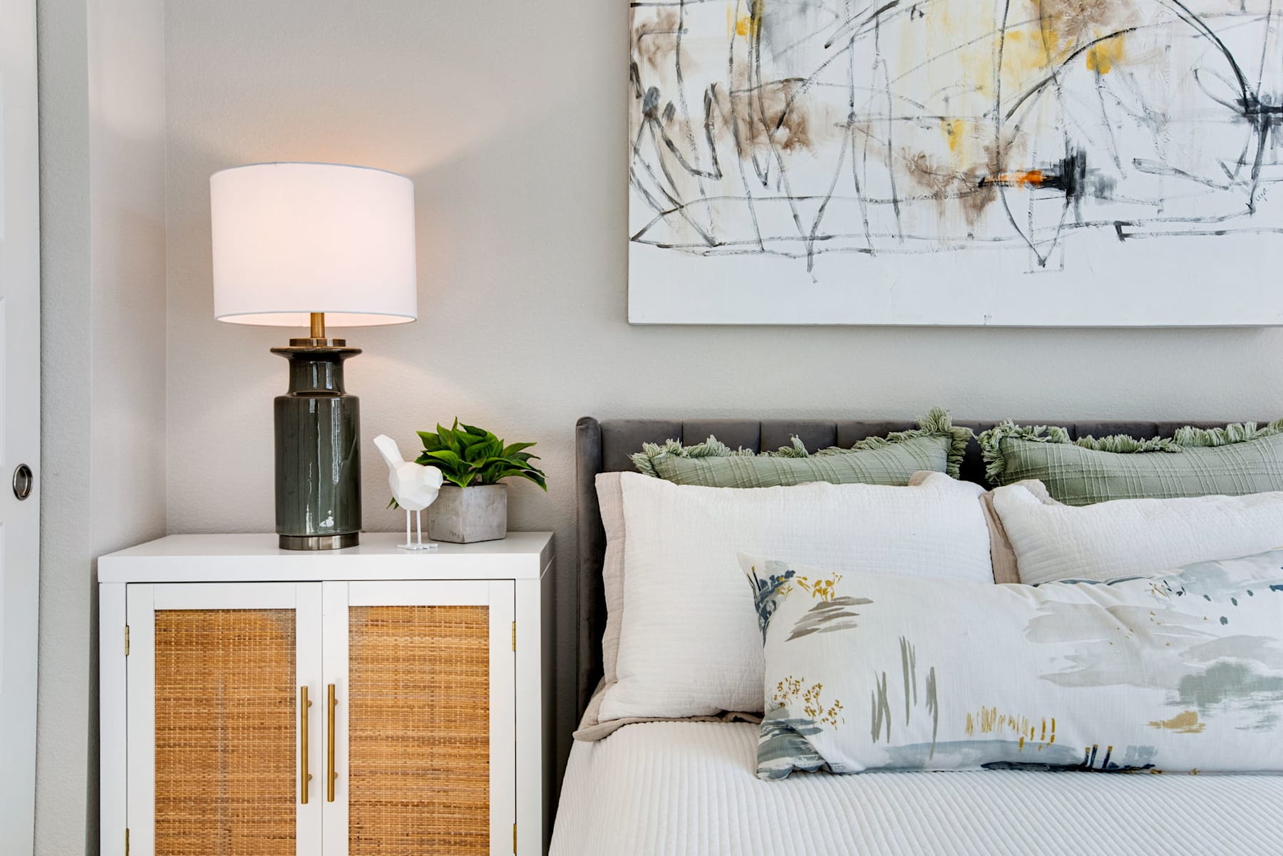 A cozy bedroom with a white bed, a lamp, and a piece of abstract art on the wall, complemented by a wooden cabinet and potted plants.