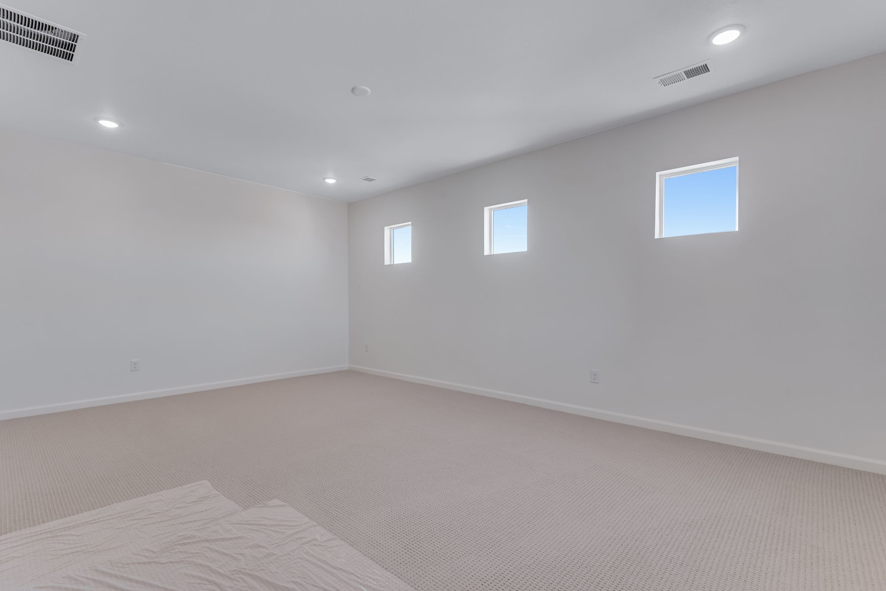 An empty, well-lit room with white walls, a carpeted floor, and several small windows providing natural light.