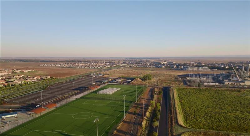 An aerial view of a suburban area with green fields, sports fields, and a railway line running through the landscape, with a city skyline visible in the distance.
