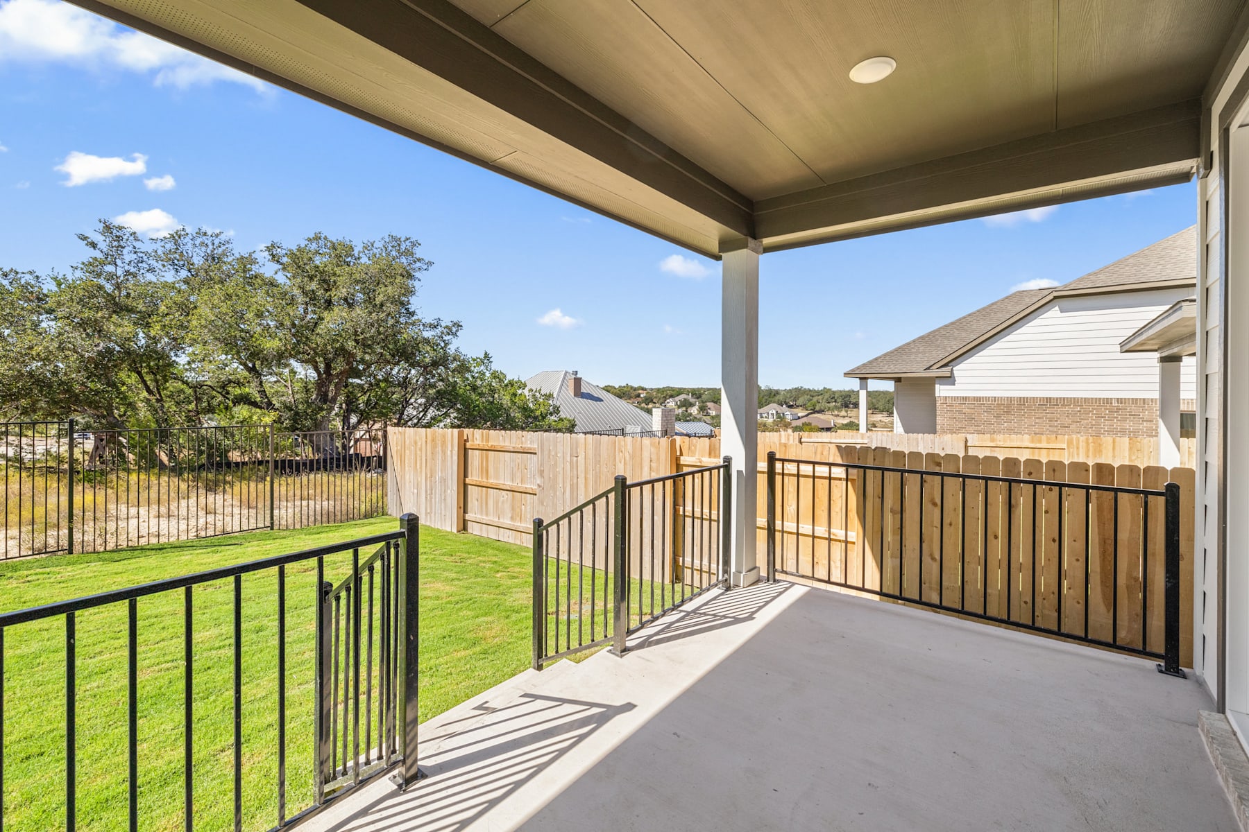 A covered patio with a metal railing overlooks a grassy backyard surrounded by trees and fencing.