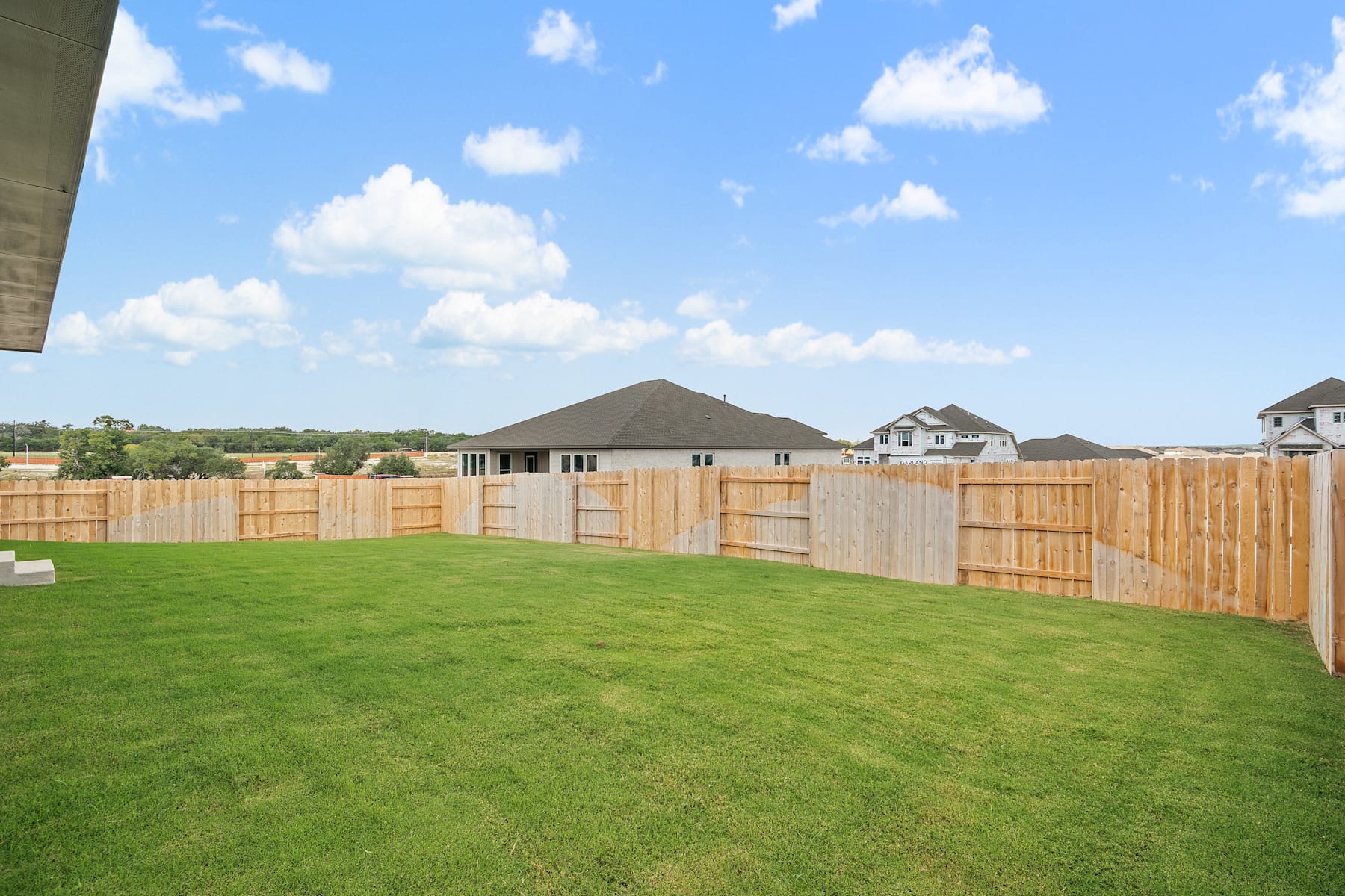 A well-manicured lawn with a wooden fence in the foreground, and a residential neighborhood with houses and a hill in the background under a blue sky with fluffy white clouds.