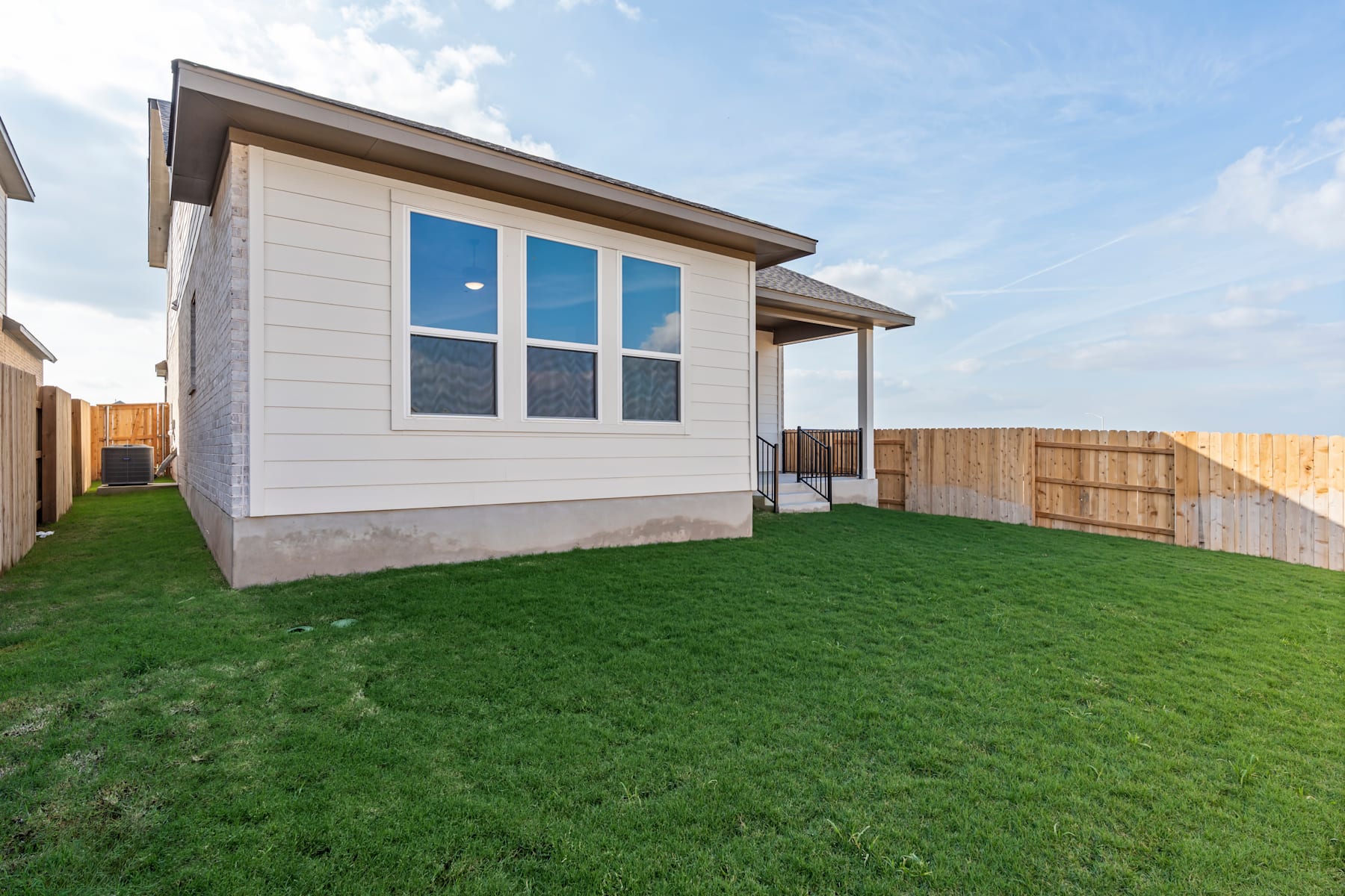 A modern, single-story house with a covered porch and large windows stands on a well-manicured lawn, surrounded by a wooden fence in the background.