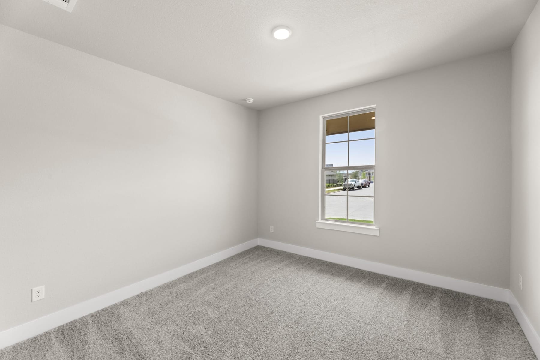 An empty room with a large window overlooking a residential area, featuring a plain white wall and gray carpeted floor.