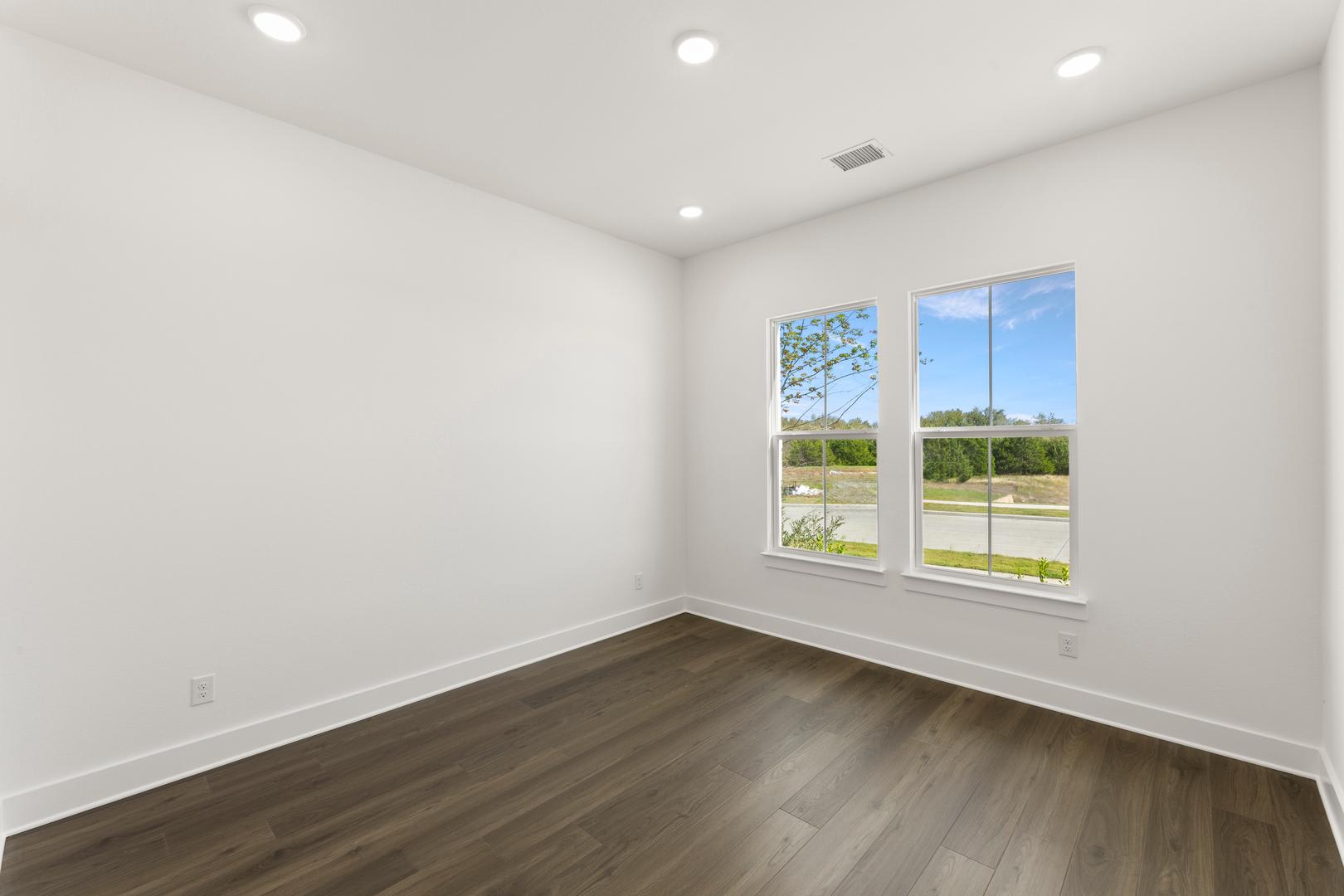 An empty room with white walls, a large window overlooking a grassy outdoor area, and a dark hardwood floor.