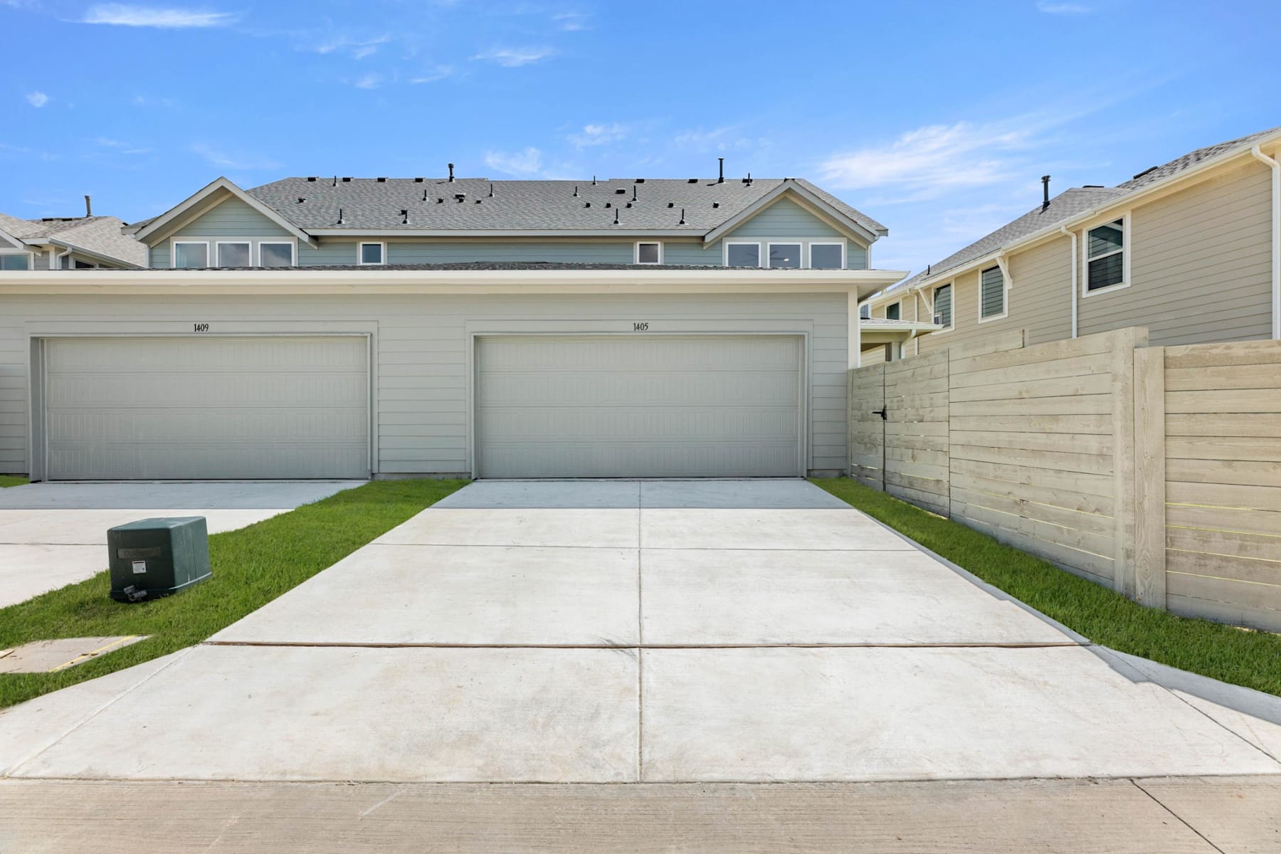 A two-story residential building with a garage, surrounded by a grassy yard and a concrete driveway, set against a clear blue sky with scattered clouds.