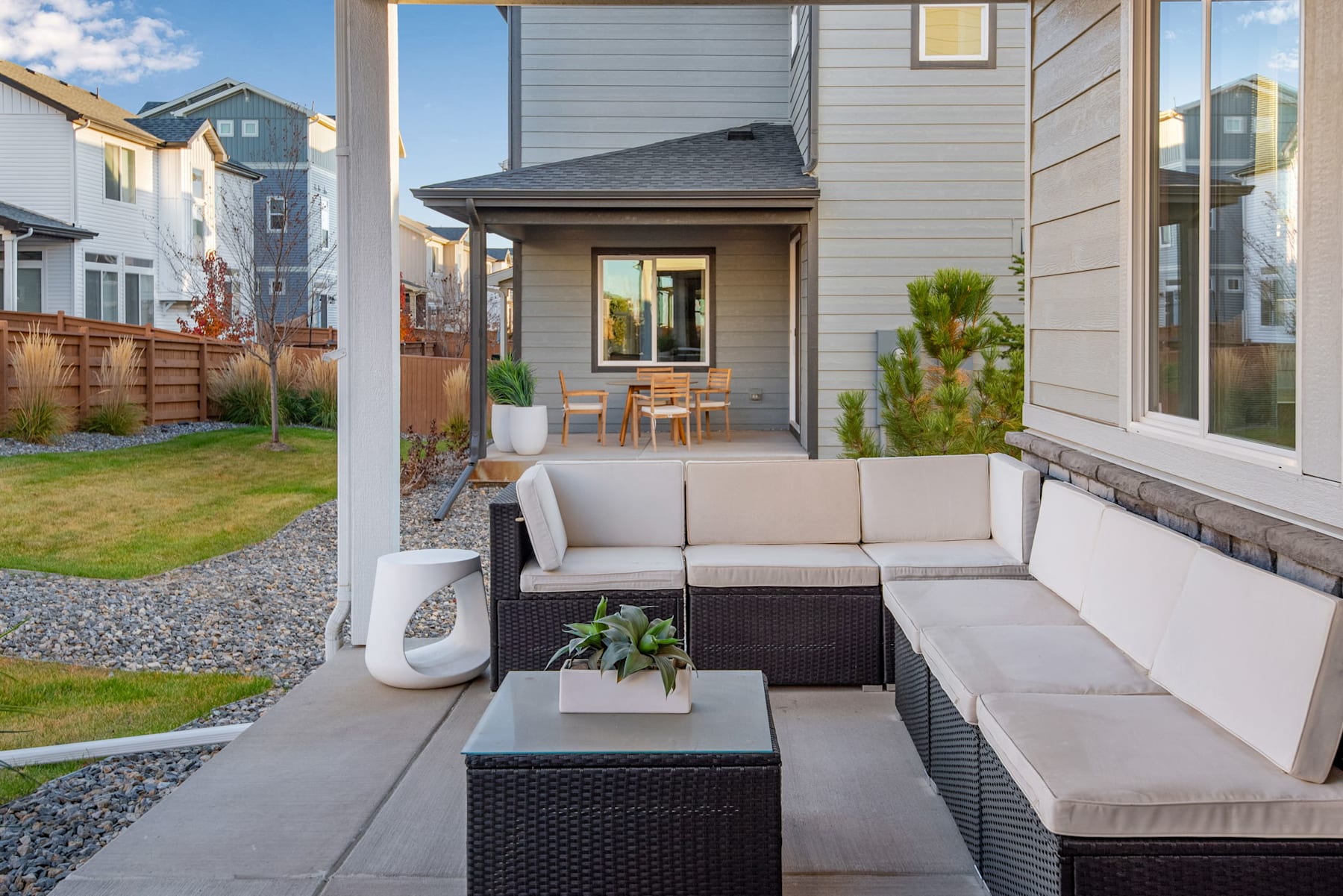 A cozy outdoor seating area with a wicker sofa and coffee table, surrounded by a well-maintained yard and a modern, two-story residential building in the background.