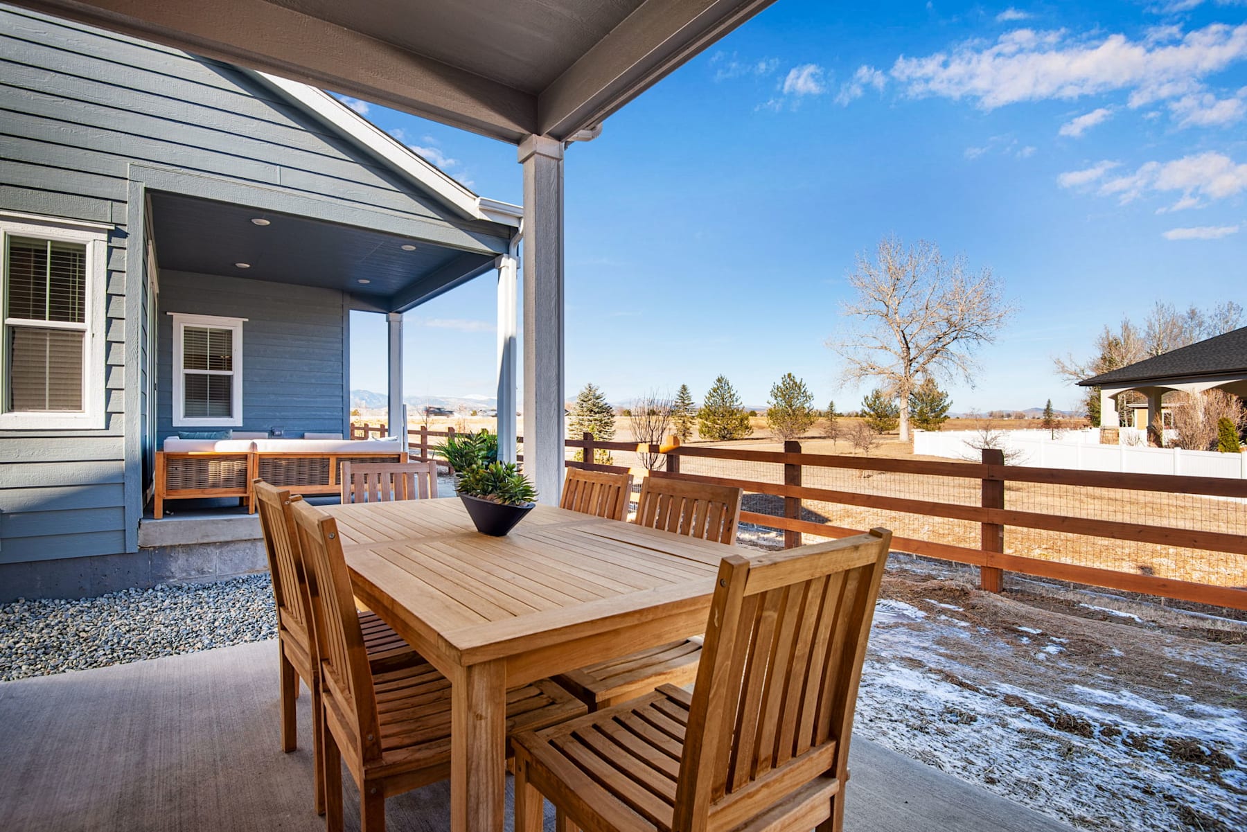 A cozy wooden patio with a dining set overlooking a snowy landscape, surrounded by a fence and trees under a clear blue sky.
