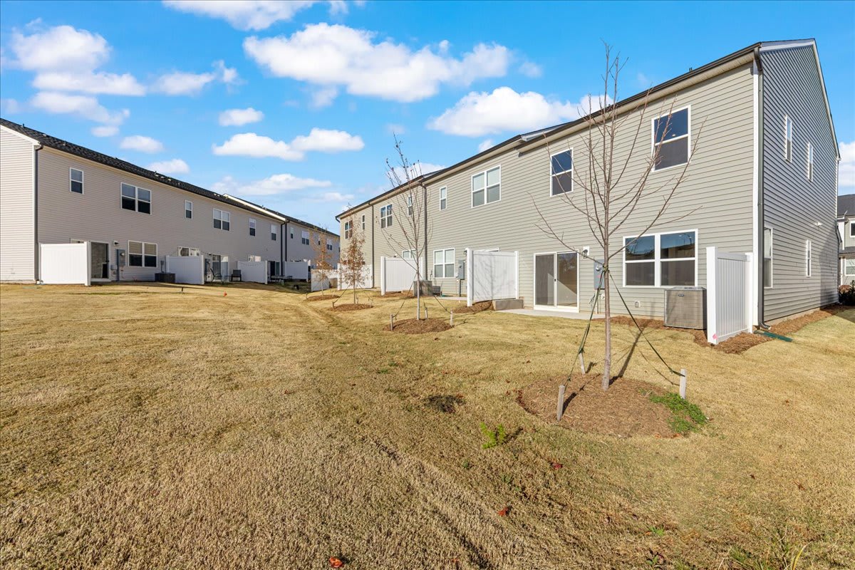A row of newly constructed townhouses with a grassy yard in the foreground, set against a backdrop of a blue sky with fluffy white clouds.