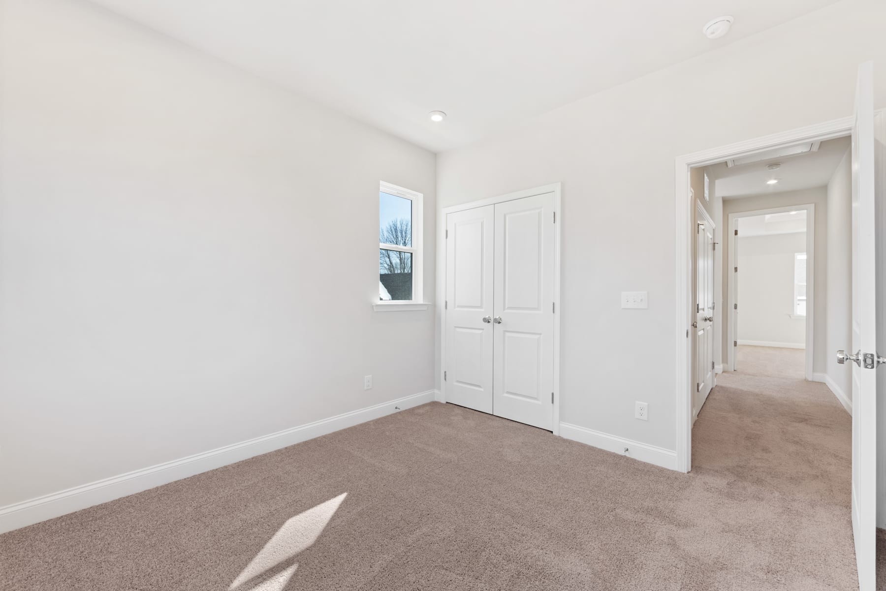 A bright, spacious room with white walls, a closet, and a carpeted floor, with a window providing natural light.