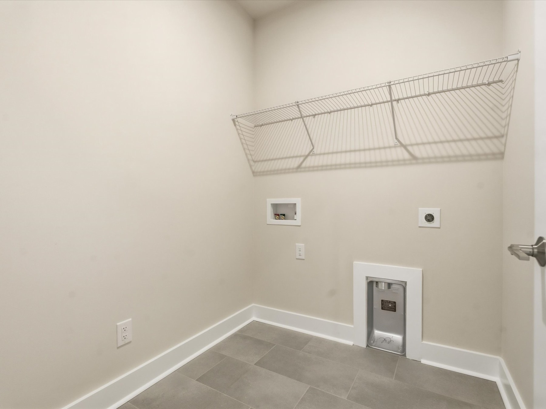 A small, empty room with white walls, a wire storage rack mounted on the wall, and a thermostat and electrical outlet visible on the wall.