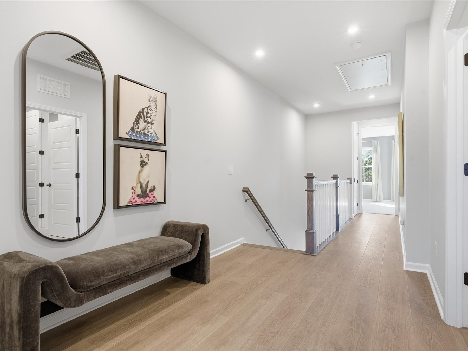 A spacious and well-lit hallway with a plush brown bench, framed artwork, and a large oval mirror adorning the walls, leading to a doorway at the end.