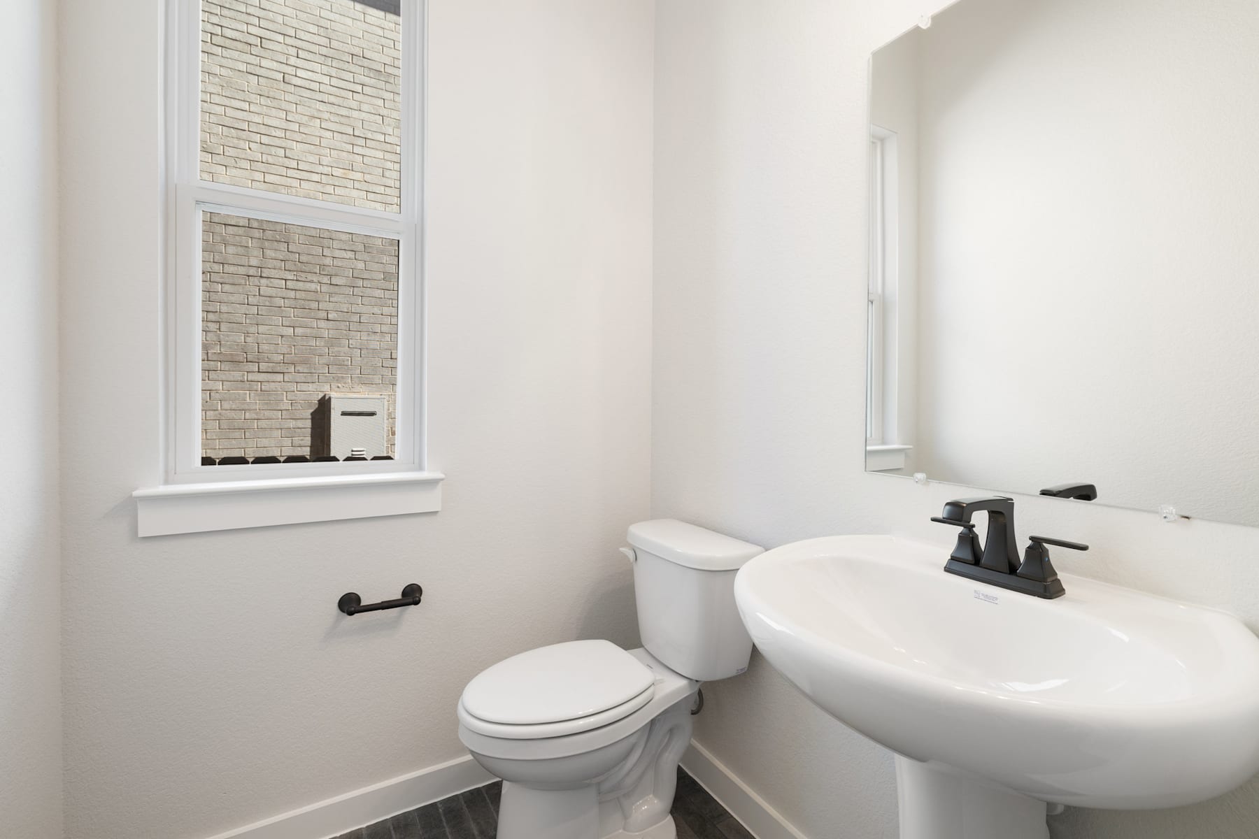 A clean, modern bathroom with a white toilet, a pedestal sink, and a window with brick accents.