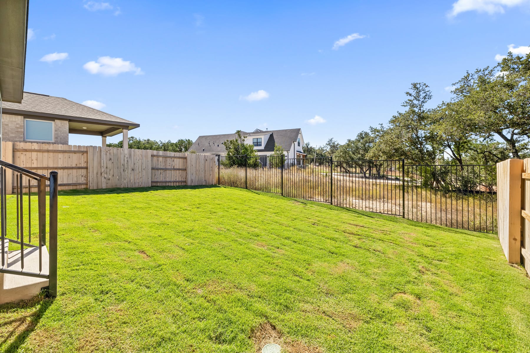 A well-manicured lawn surrounded by a wooden fence, with a porch and trees visible in the background against a blue sky with fluffy clouds.