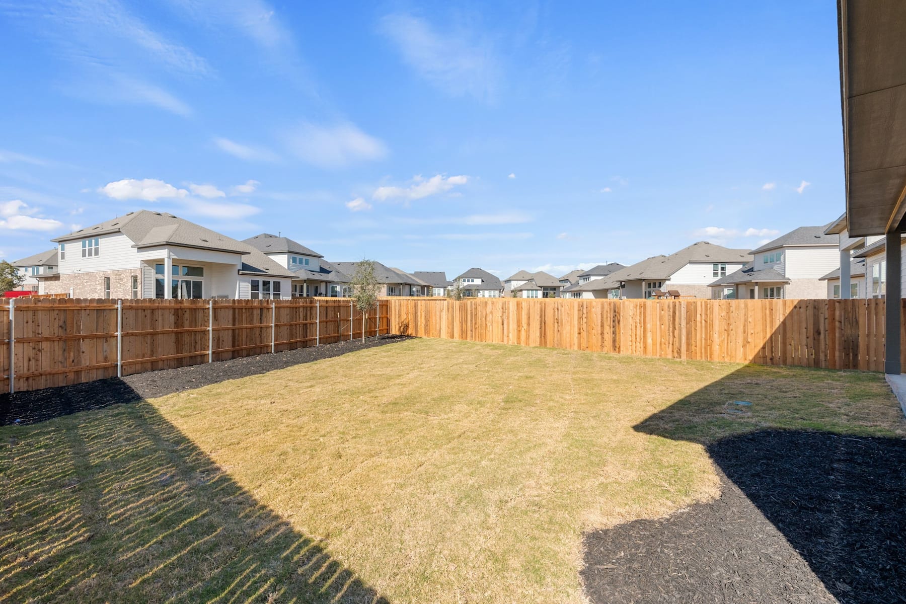 A large grassy backyard surrounded by wooden fences, with several houses visible in the background under a clear blue sky.