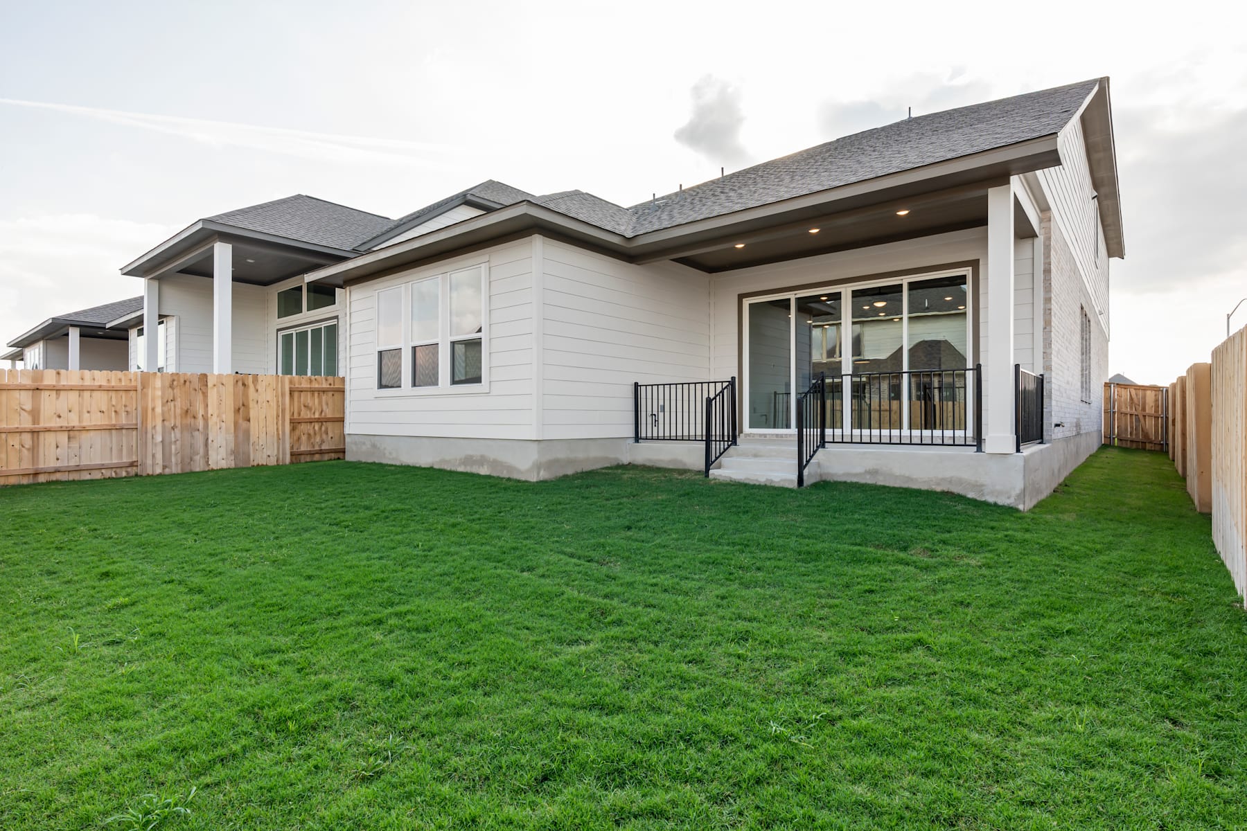A modern, two-story residential house with a well-manicured lawn in the foreground, surrounded by a wooden fence and set against a clear sky in the background.