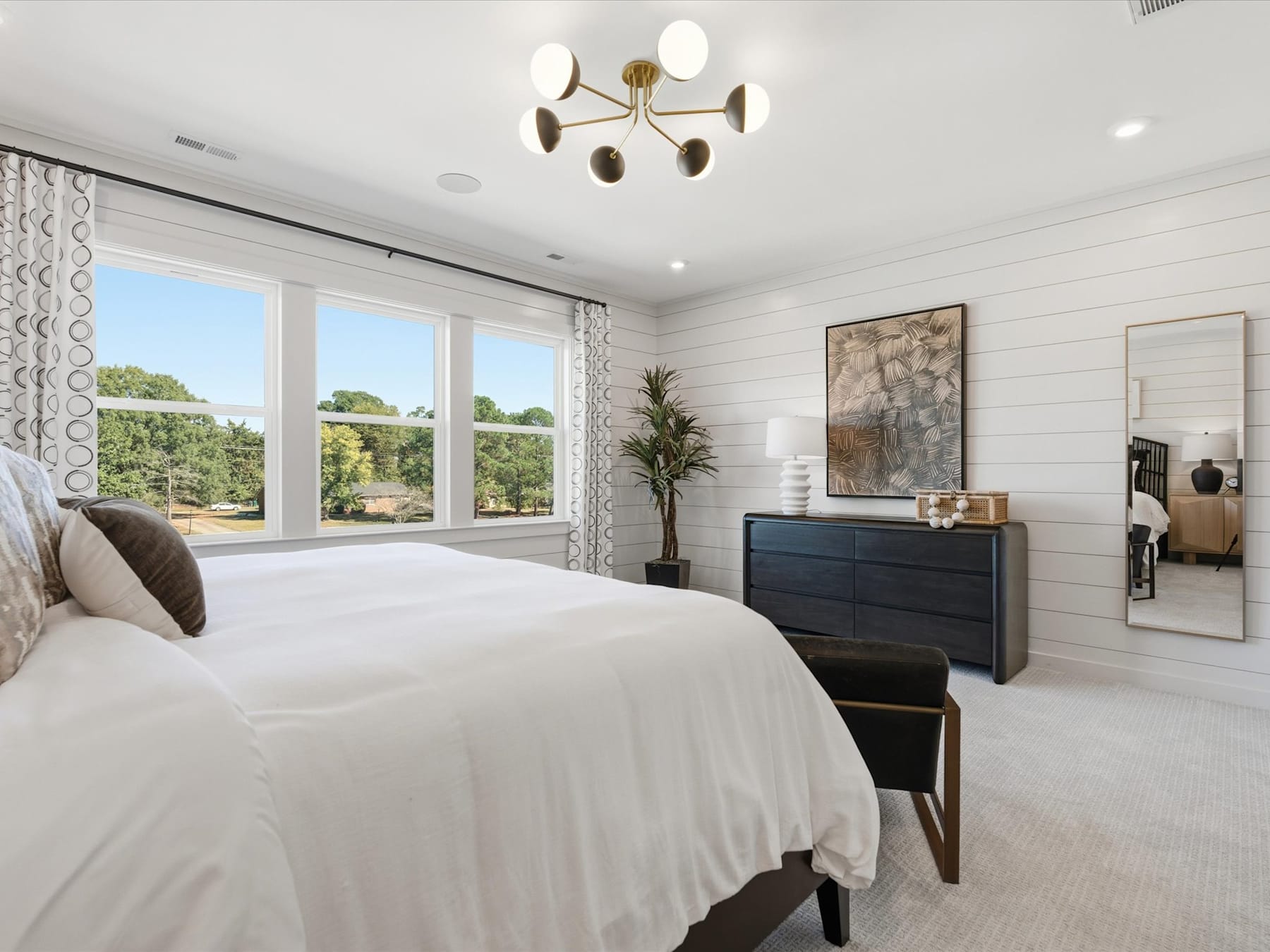 A spacious and well-lit bedroom with a large window overlooking a scenic outdoor landscape, featuring a modern chandelier, a dresser, and various decorative elements.