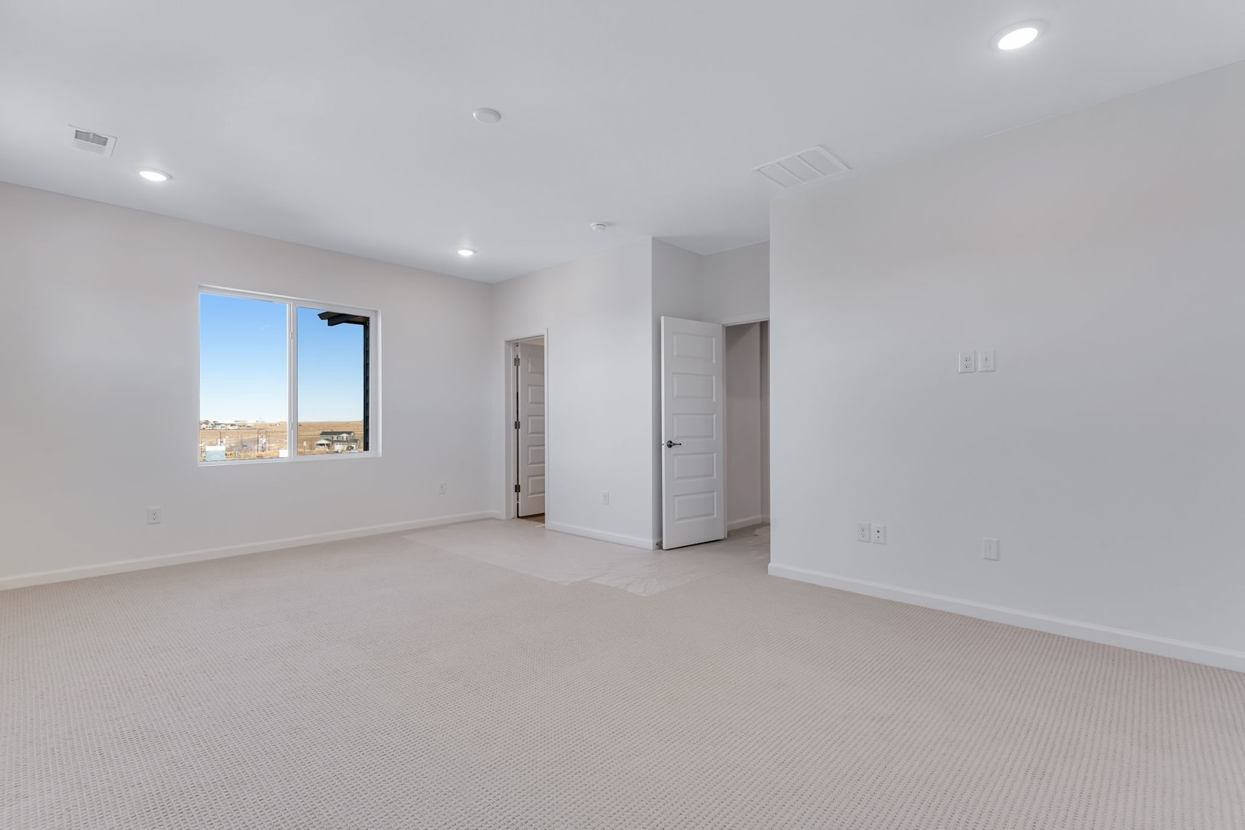A spacious, well-lit room with white walls, a window, and a carpeted floor. The room appears to be a bedroom or living space with a clean, minimalist design.