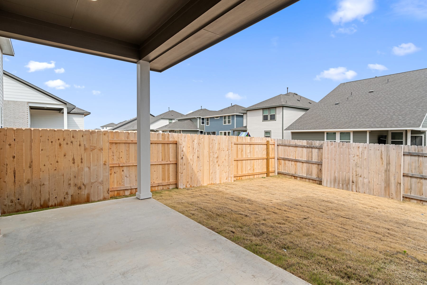 A covered patio area with a wooden fence in the foreground, surrounded by residential homes in the background under a blue sky with clouds.