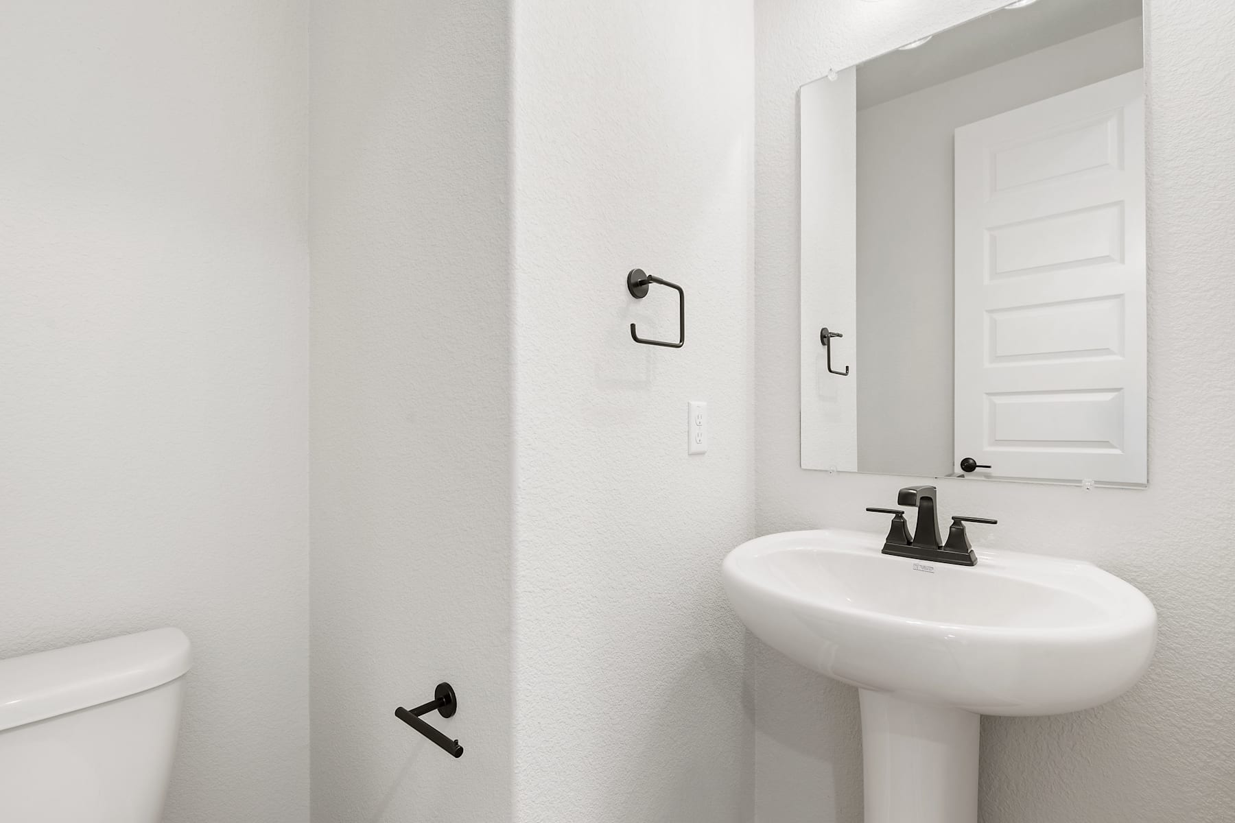 A simple, minimalist bathroom with a white pedestal sink, a mirror, and a white toilet against a plain white wall.