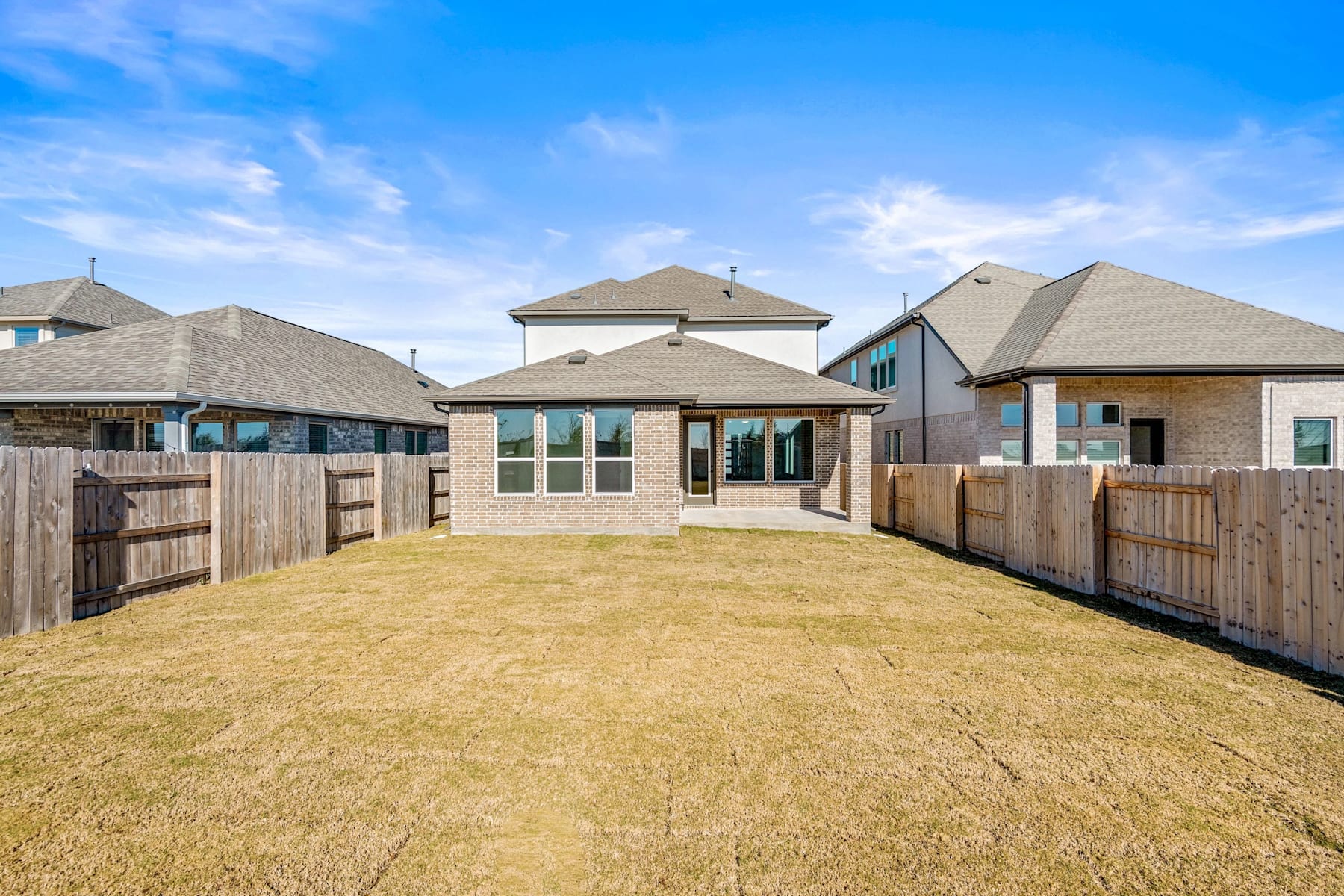 A well-manicured lawn leads to a row of modern, two-story houses with tan roofs and siding, set against a bright blue sky with scattered clouds.