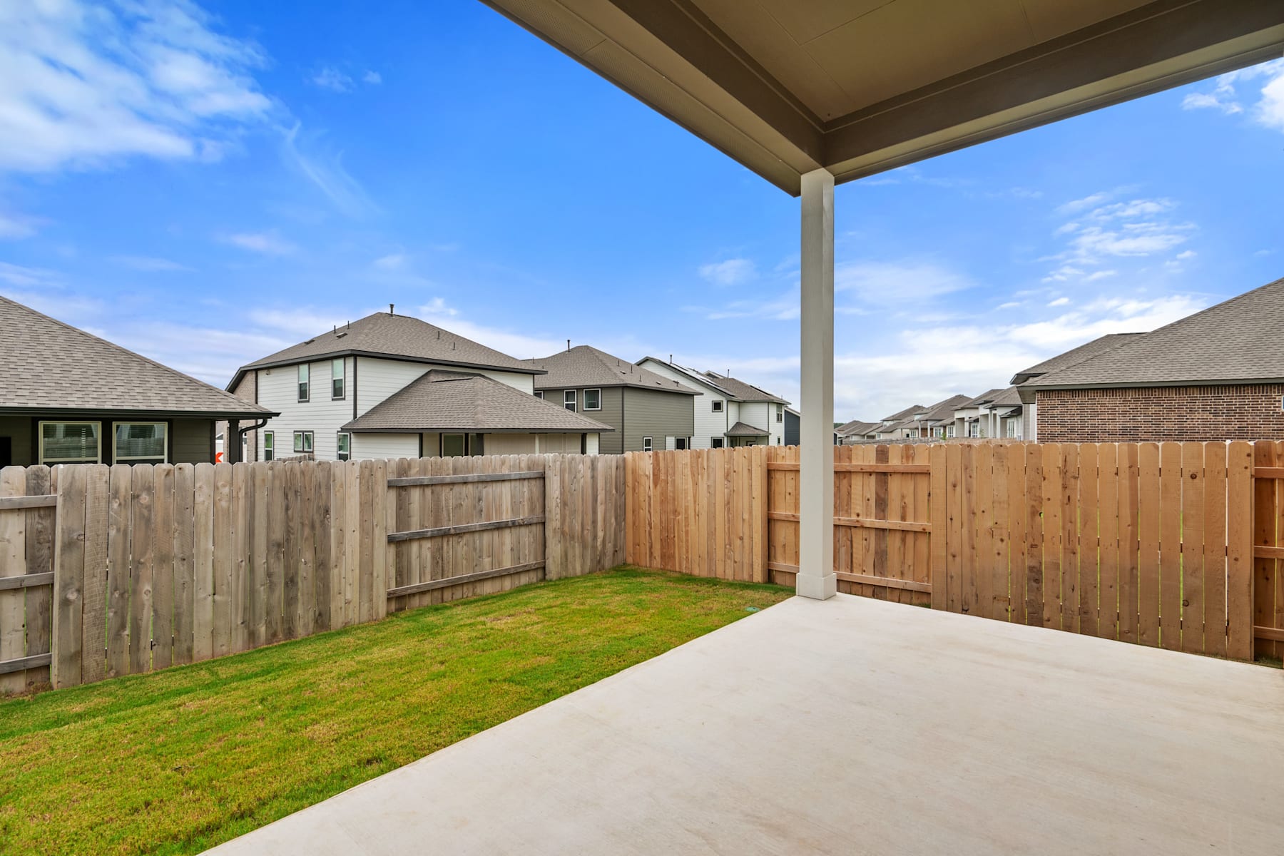 A covered patio with a wooden fence and a grassy yard, surrounded by residential houses in the background under a blue sky with clouds.