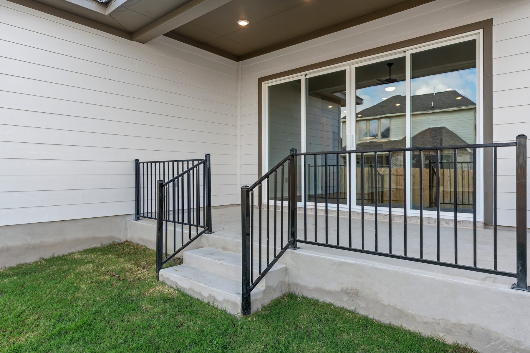 A covered porch with a metal railing leads to a grassy yard, with a residential building visible in the background through the glass doors.